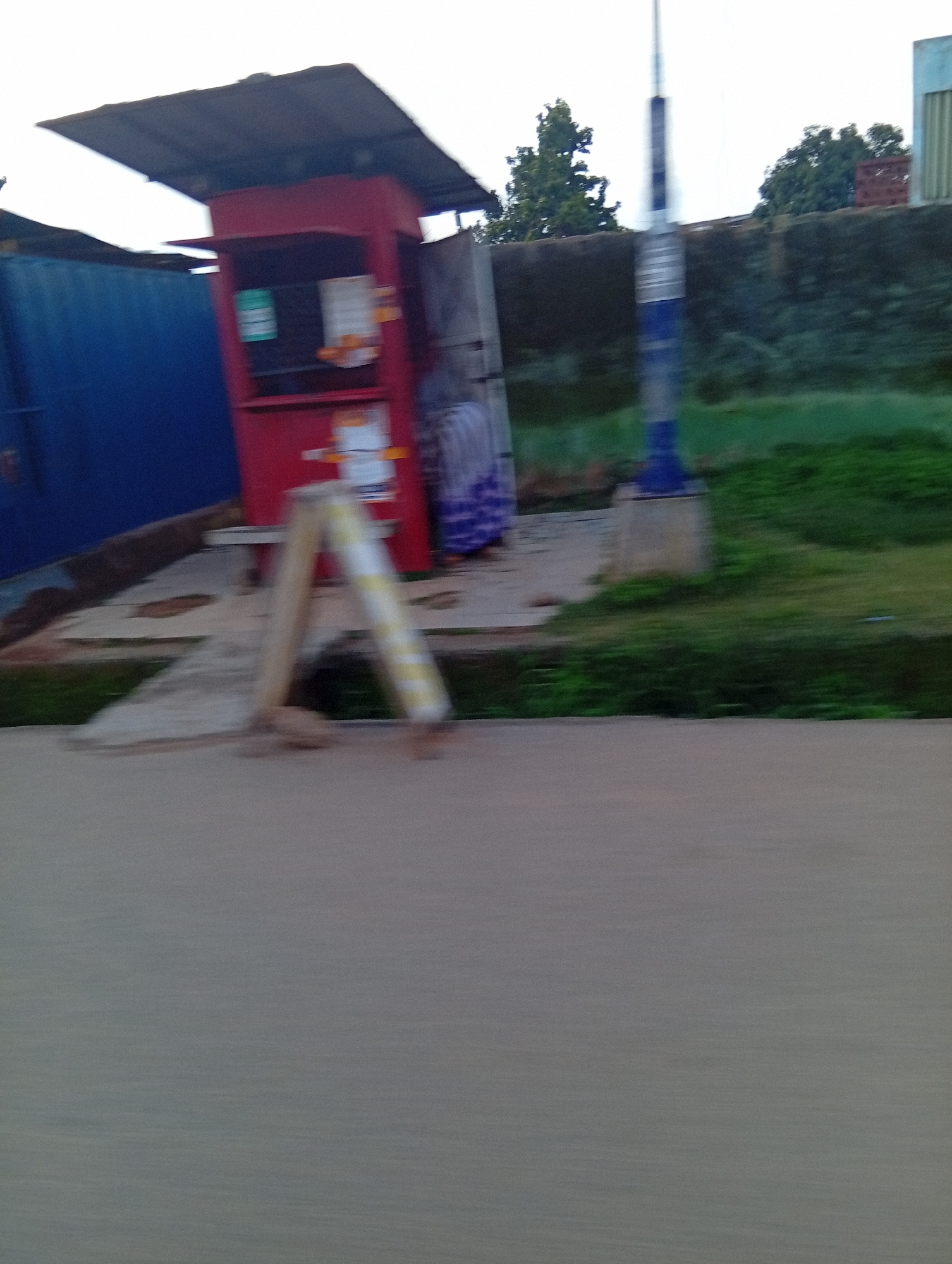 The image shows a blurry, outdoor scene in what appears to be a city. On the left, a large, blue container-like structure is partially visible. Adjacent to it is a small, red kiosk with a dark-colored roof. The kiosk has openings, and what appear to be signs or posters are visible inside. In front of the kiosk, an A-frame sign with yellow and white stripes is propped up. A person wearing a purple and white patterned garment is partially visible behind the kiosk. The ground in front of the kiosk appears to be paved with concrete. To the right of the kiosk, a blurry, grassy area with some trees and a wall can be seen. A tall, blue and grey pole with visible bands stands in the middle ground. The scene is captured with motion blur, suggesting movement of the camera or the subjects. The overall impression is that of a street-side scene, possibly a market or a small roadside business. Based on the provided metadata, the location is identified as Jos, Nigeria.