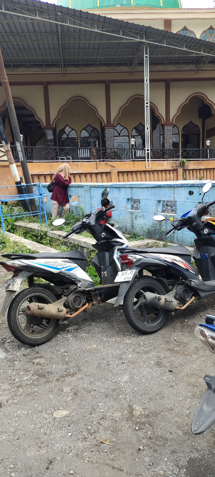 The image shows two motorcycles parked in front of a building in Kecamatan Lawang, Indonesia. The motorcycles are parked on a gravel surface. The building is a mosque with a green dome. The motorcycles are a black and white Honda Vario 150 and a black and red Honda Beat. The image was taken on a cloudy day. A woman in a brown hijab and beige pants is visible in the background walking towards the mosque. A portion of the mosque's roof is covered with a metal awning. The mosque's exterior wall is a light blue color. The motorcycles are facing the mosque.