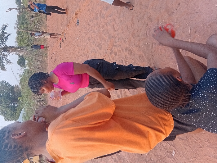 Three people are outside, possibly on a sandy area. One person wears an orange shirt and faces away from the camera. A person in a pink shirt drinks from a clear cup. Another person is to the right in a navy blue polka dot shirt, holding an orange item in their hands. Other people can be seen in the background, including someone wearing a blue shirt and black pants. There is a tree in the background.