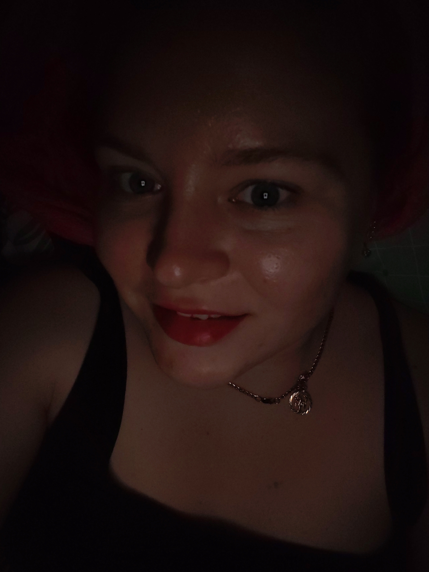 This media file captures a close-up, low-light selfie of a person, likely female, illuminated primarily from below, suggesting light from a screen. The subject has fair skin, a round face, and red hair visible around the edges of the frame.
The person is looking directly at the camera with wide, dark eyes. Bright, rectangular reflections, consistent with light from a screen or digital display, are clearly visible in the pupils of both eyes. A subtle, closed-mouth smile is present, highlighted by vibrant red lipstick. The skin on the forehead, nose, and cheeks appears somewhat shiny, a common effect of direct, close-up lighting.
The subject is wearing a dark-colored, possibly black, tank top or camisole. A delicate, gold-toned necklace with a circular pendant adorns the neck. A small, metallic earring is also visible in the right ear.
The background is predominantly dark and out of focus, indicating an indoor setting with minimal ambient light. A faint green grid pattern, possibly part of a mat or craft surface, can be discerned behind the right side of the person's head. The overall impression is one of direct engagement, characteristic of a personal portrait taken in a dim environment, possibly at night. No visible text is present in the image. The context for this image is Izhevsk, Russia.