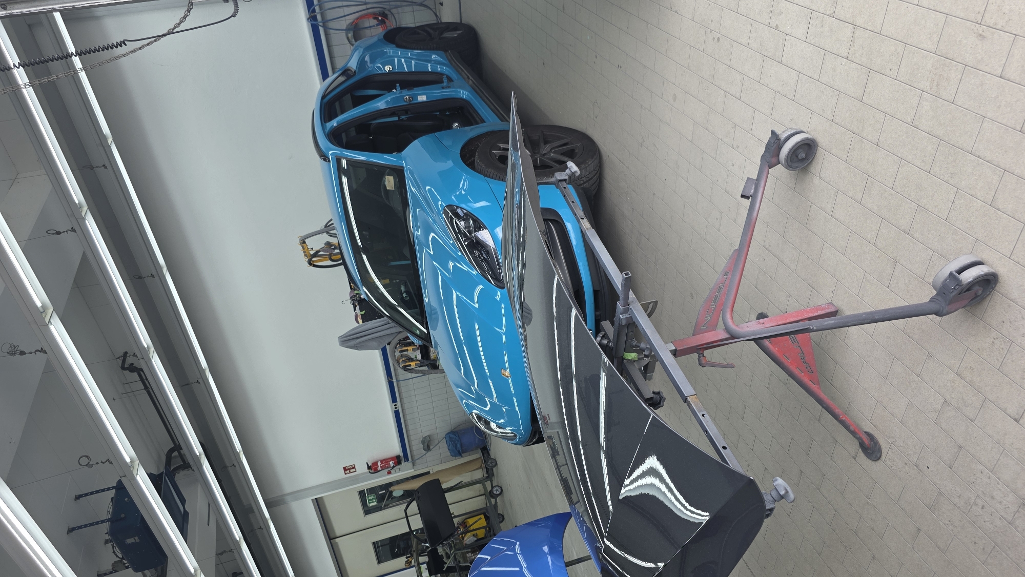 In a well-lit, tiled auto repair shop, a bright blue Porsche Macan is being worked on. The car's doors are open, and the hood is detached and placed on a red, wheeled stand in the foreground. The hood is dark and highly reflective, showing the overhead lights. Various tools and equipment, including a fire extinguisher and a blue car panel, are visible in the background. The overall scene suggests an ongoing automotive repair or restoration process.