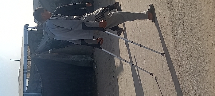 A person using crutches stands outside. They wear gray sweatpants, a gray sweatshirt, and sandals. Another person is partially visible in the background, wearing dark clothing. To the left, a gray structure or building is visible. The ground appears to be concrete. The person with crutches casts a long shadow, suggesting bright sunlight.