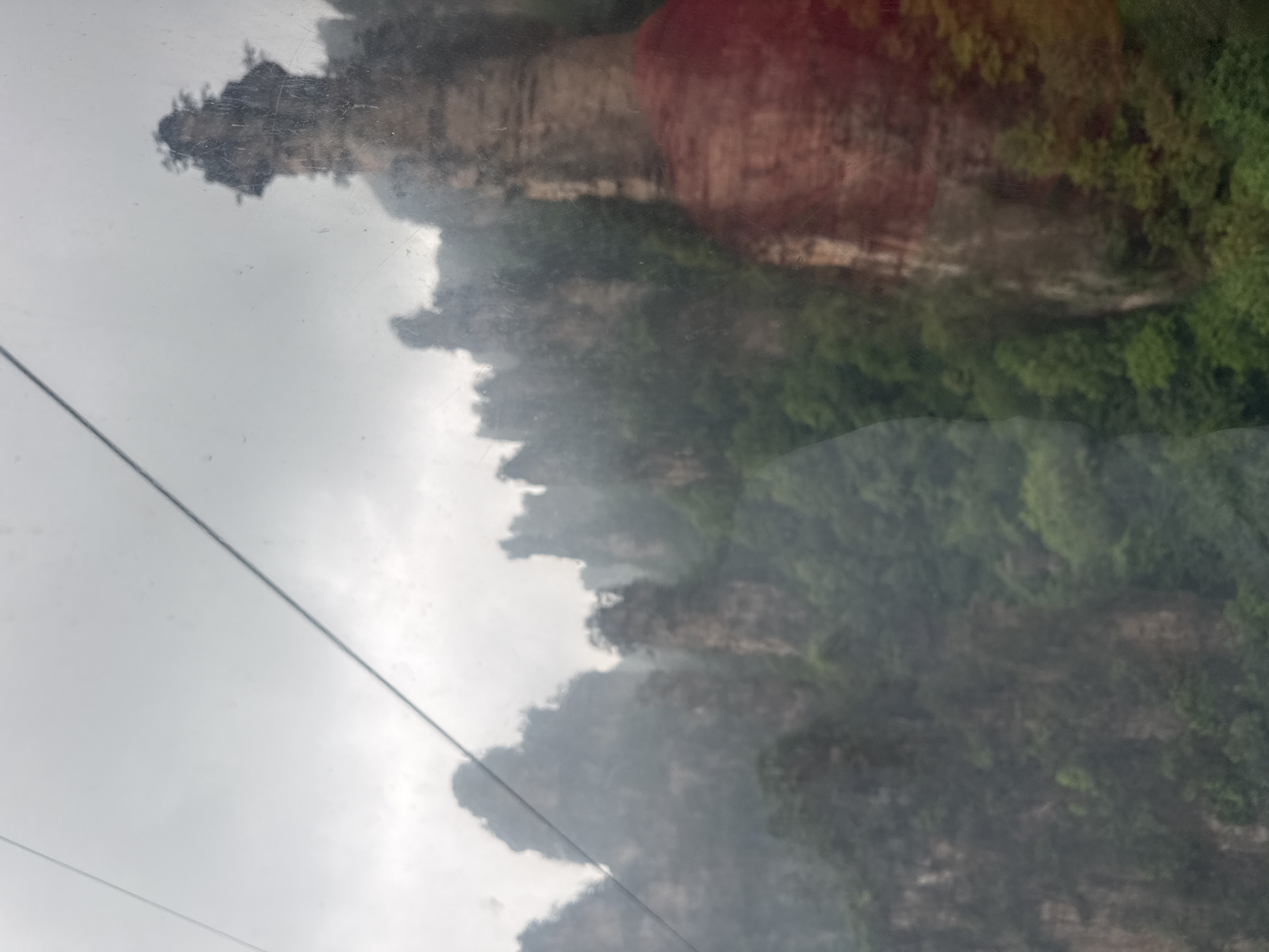 The image shows a landscape, possibly viewed through a window, featuring a mountain range with unique rock formations and lush vegetation. The sky is overcast, and there's a cable or wire running diagonally across the left side of the frame. The overall scene is somewhat obscured by mist or fog. The location is consistent with the Wulingyuan Scenic and Historic Interest Area in China, known for its sandstone pillars.