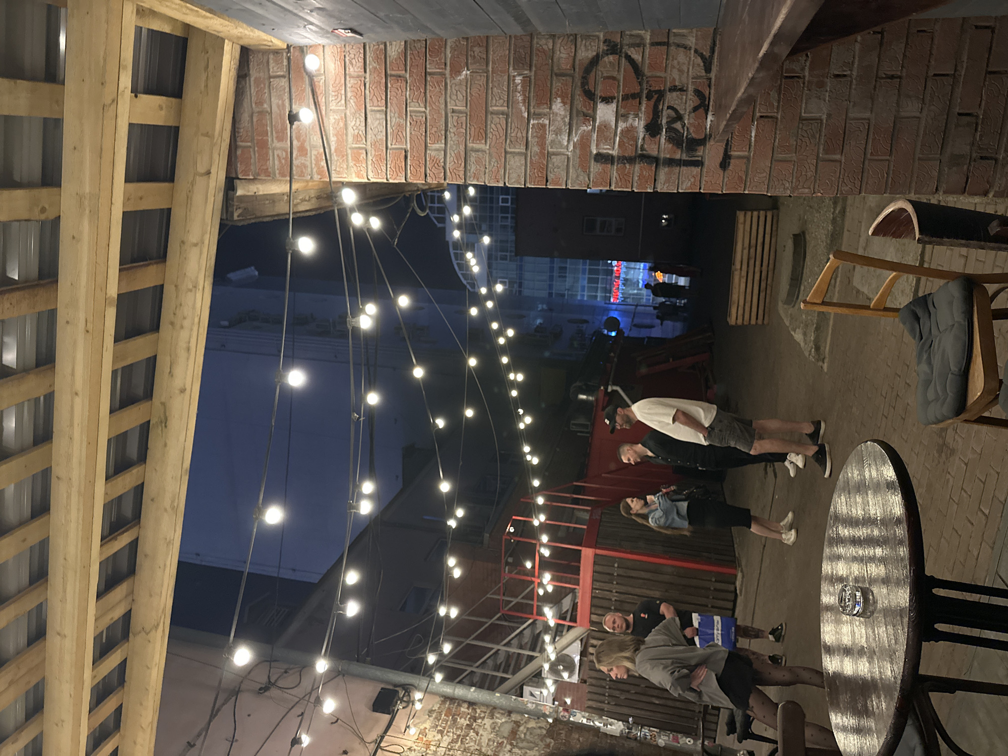 Here is a description of the image:
**Location:** The image appears to be taken in an outdoor courtyard or alleyway. While the prompt mentions Perm, Russia, there are no identifying characteristics in the image itself to confirm this location.
**Environment:** The courtyard is partially enclosed by brick buildings and wooden structures. A string of light bulbs stretches across the courtyard, providing illumination. The ground is paved with bricks. There's graffiti on the brick wall.
**People:** There are several people visible in the scene. They appear to be standing and talking to each other. The subjects are positioned in the center of the photograph.
**Objects:** A round table is visible in the foreground. A chair is near the table. There is what appears to be a pallet in the background. A set of metal scaffolding with red railings.