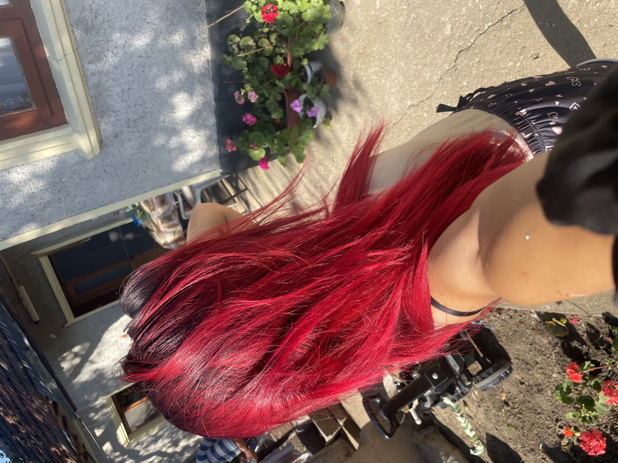 In Zheglartsi, Bulgaria, a person with vibrant, bright red hair is captured from a rear, high-angle perspective, standing in a sunlit, outdoor courtyard. The person's hair is the dominant feature, cascading down their back, with visible roots showing darker, natural hair color. They are wearing black shorts adorned with small pink hearts and a black bra strap is visible.
The environment is rustic, with a weathered building in the background featuring a brown-framed window and a dark-tiled roof. The building's exterior is light-colored and textured. Shadows from trees are cast across the ground, indicating a sunny day. In the foreground, to the left, a black wheeled device, possibly a garden cart or a trolley, rests on a patch of soil. Several potted plants with pink and red flowers are arranged along the base of the building. The ground is a mix of concrete and dirt, with some cracks visible in the concrete. The overall impression is a casual, outdoor setting during daylight hours.