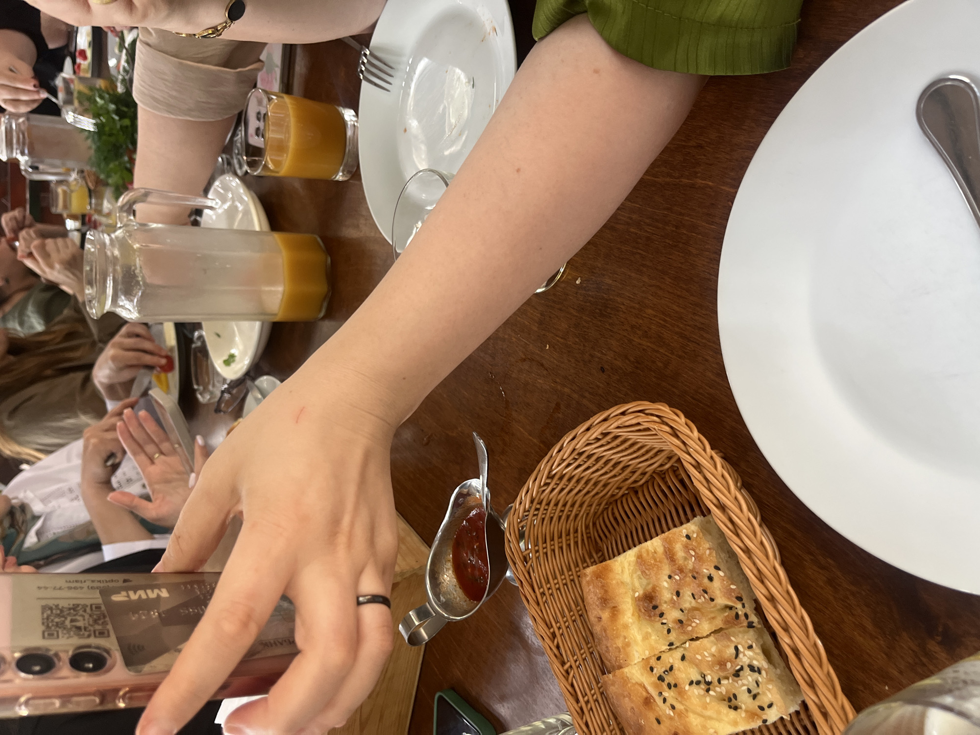 The image depicts a group of people seated at a wooden table, engaged in a meal. In the foreground, a person's arm is visible, holding a smartphone that has a payment card inserted into its case. A small metal dish containing a red sauce, likely ketchup or a similar condiment, is placed next to a woven basket holding two pieces of flatbread sprinkled with sesame and black seeds. Further back, other individuals are visible, with plates and glasses on the table. A glass pitcher filled with orange juice and a glass of the same beverage are also present. The background shows hints of other diners and their meals. The scene suggests a casual dining experience in a restaurant.