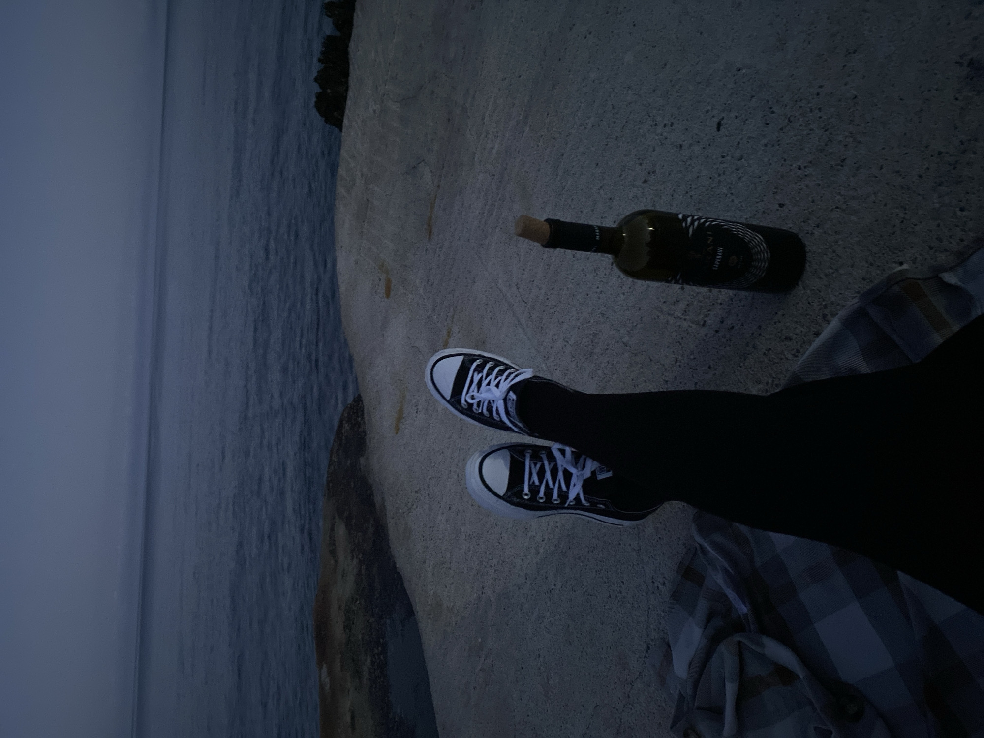 A person wearing black leggings and black and white Converse sneakers sits on a concrete surface next to the Adriatic Sea in Kunje, Montenegro. A partially consumed bottle of wine with a cork lies nearby. The person is resting on a plaid blanket. The environment suggests either dusk or dawn due to the dim lighting and low visibility over the water.