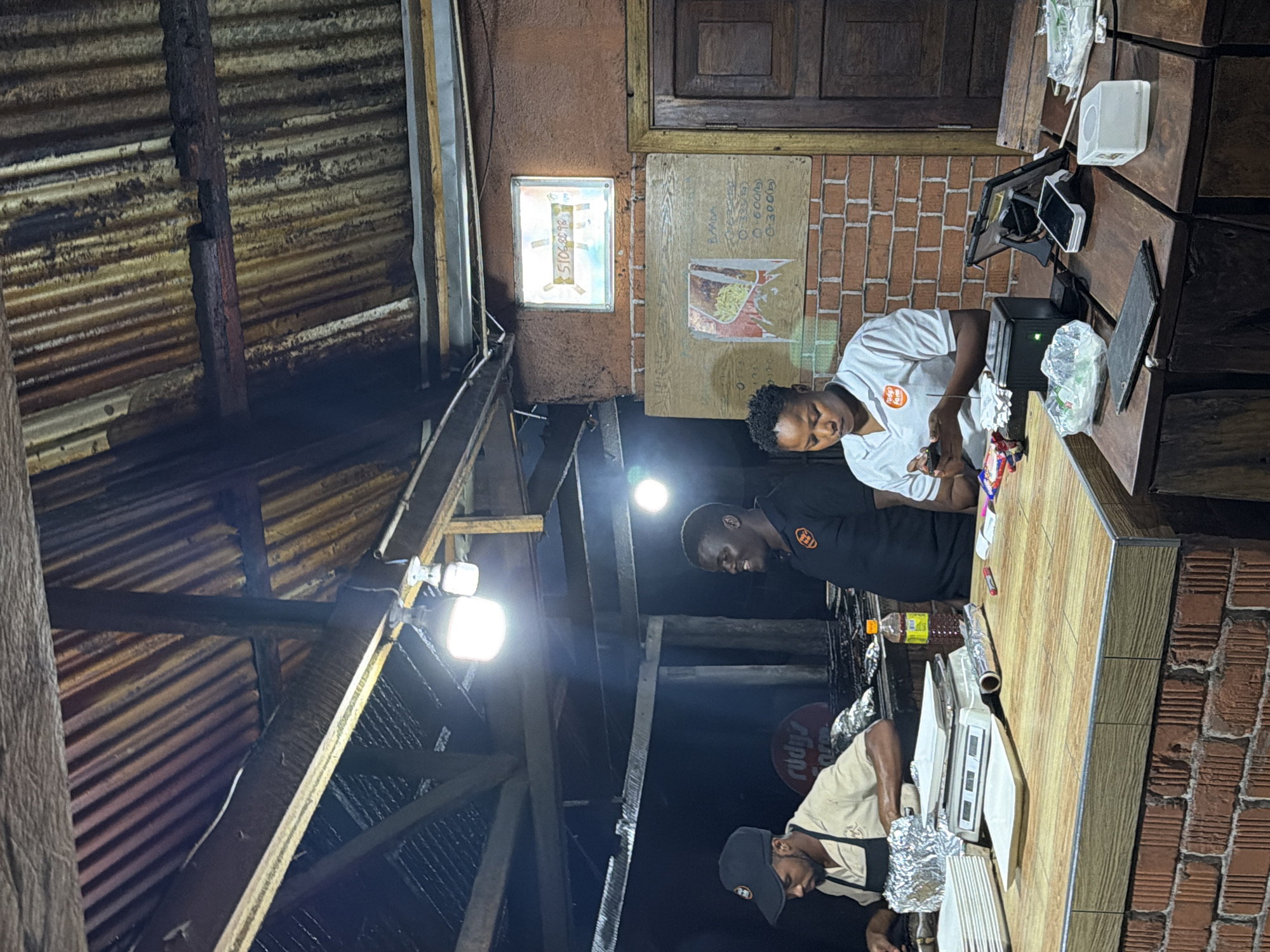 The image shows a scene inside a food stall, likely in Dar es Salaam, Tanzania, characterized by its rustic construction. A corrugated metal roof, supported by wooden beams, is visible overhead. Three individuals appear to be working at a counter. On the left, a man wearing a black cap and apron is preparing food, with foil-wrapped items and stacks of plates on the counter. To his right, another man in a black polo shirt with an orange logo is smiling and looking to his left. In the center, a woman in a white polo shirt with an orange logo is looking down at her phone and holding a device. The counter displays a digital scale, a bottle of what appears to be cooking oil, and various electronic devices, including a tablet on a stand and card readers, suggesting a commercial transaction is occurring. The background reveals a brick wall with a framed digital display showing a phone number and a wooden door. The overall atmosphere suggests a busy, open-air food service environment.