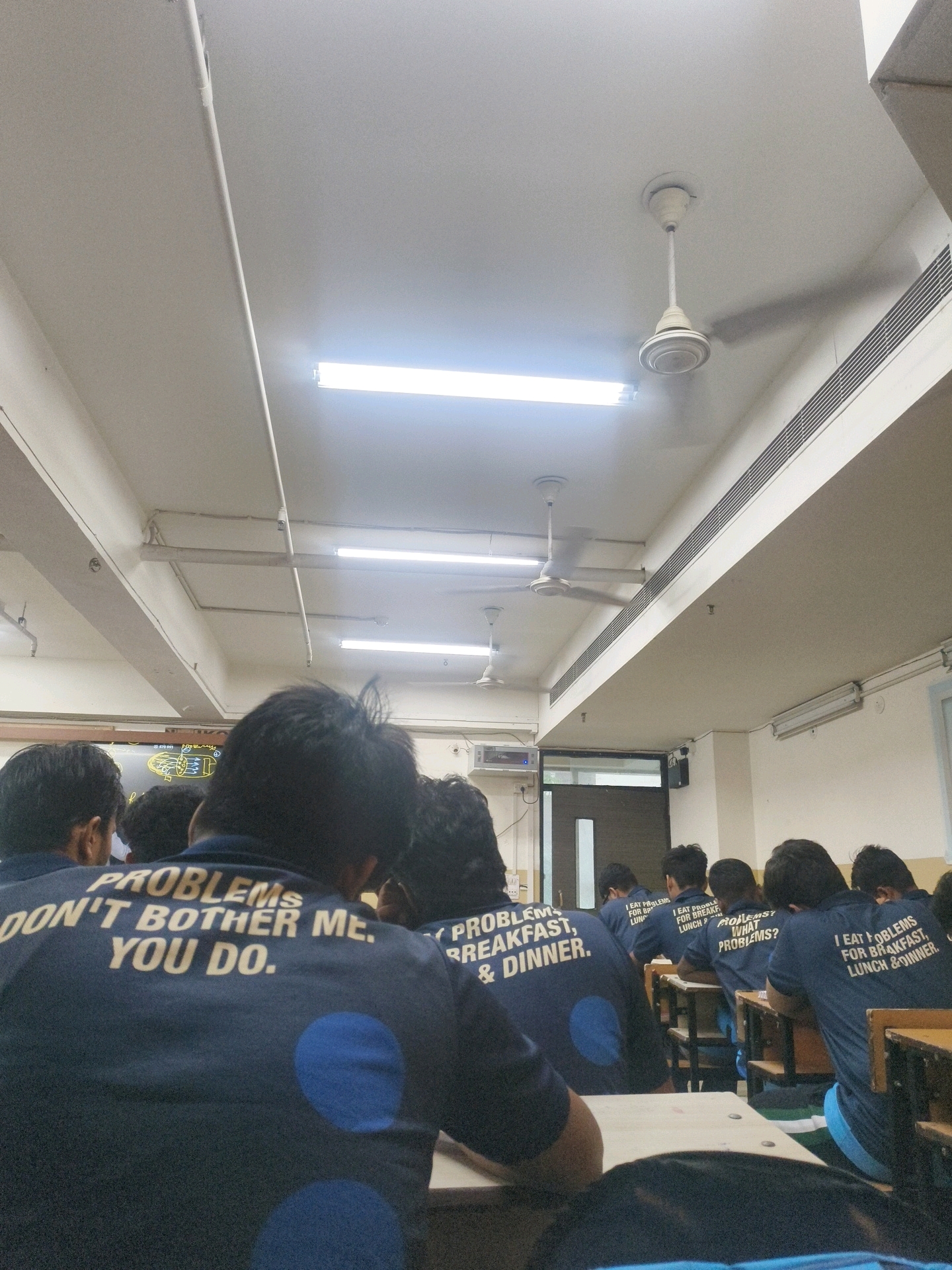The image captures an indoor scene, likely a classroom or lecture hall, in Kota, India. Several young individuals, presumably students, are seated at desks facing away from the camera. They are all wearing navy blue t-shirts with white text printed on the back. The text on the t-shirts reads, "PROBLEMS DON'T BOTHER ME. YOU DO." and "I EAT PROBLEMS FOR BREAKFAST, LUNCH & DINNER." The environment appears to be functional, with fluorescent lighting panels and ceiling fans visible on the white ceiling. The students are seated at wooden desks, suggesting an educational setting. The overall atmosphere suggests a casual academic environment, possibly a break or a lecture where students are relaxed.