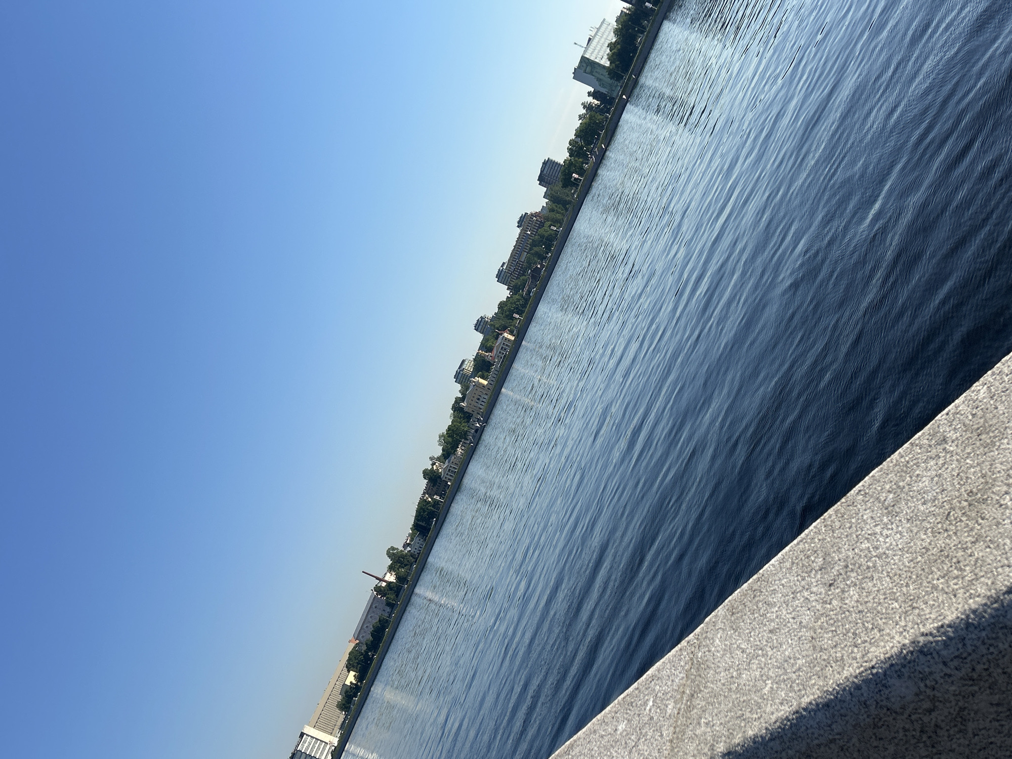 The image presents a serene urban landscape dominated by a wide body of water, likely a river or canal, under a vast, clear blue sky. The sky transitions subtly from a deeper blue overhead to a lighter shade near the horizon. Along the opposite bank of the water, a cityscape unfolds, featuring a mix of buildings, some appearing modern and others older, possibly with industrial elements. Dense green foliage from trees lines the bank, interspersed between the structures. A distinct, tall, red-brick industrial chimney or smokestack is visible on the left side of the distant bank.
The water, a deep blue hue, shows gentle ripples across its surface, reflecting the bright sunlight and creating a shimmering texture. In the immediate foreground, to the right, a textured light grey stone embankment or railing occupies the corner of the frame. A strong shadow is cast upon this stone surface, indicating bright sunlight and a clear day. No people or specific activities are visible within the frame, emphasizing the quiet stillness of the scene. The overall impression is that of a clear, sunny day, likely morning or afternoon, given the quality of light and shadows. There is no visible text in the image.