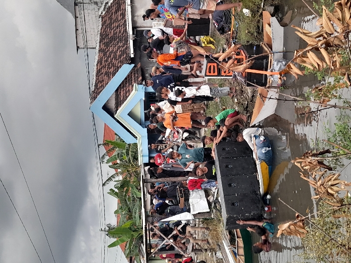 The image shows a scene of flooding in Kecamatan Tanggulangin, Indonesia, specifically at the coordinates -7.504 latitude and 112.746 longitude. There are numerous individuals, mostly men, standing, walking, and interacting in the flooded area. Some are carrying objects, others are looking at the floodwater, while some appear to be helping to move objects. There are also children in the scene. Some of the people are standing in front of a two-story house with a blue roof. Other objects in the scene include boats, large speakers, and other debris, such as chairs and pieces of wood, floating in the water. The water appears to be relatively shallow and brownish-grey in color. The sky is overcast with grey clouds. The environment is characterized by trees and foliage, suggesting a suburban or rural setting. The image conveys a sense of chaos and disruption caused by the flood.