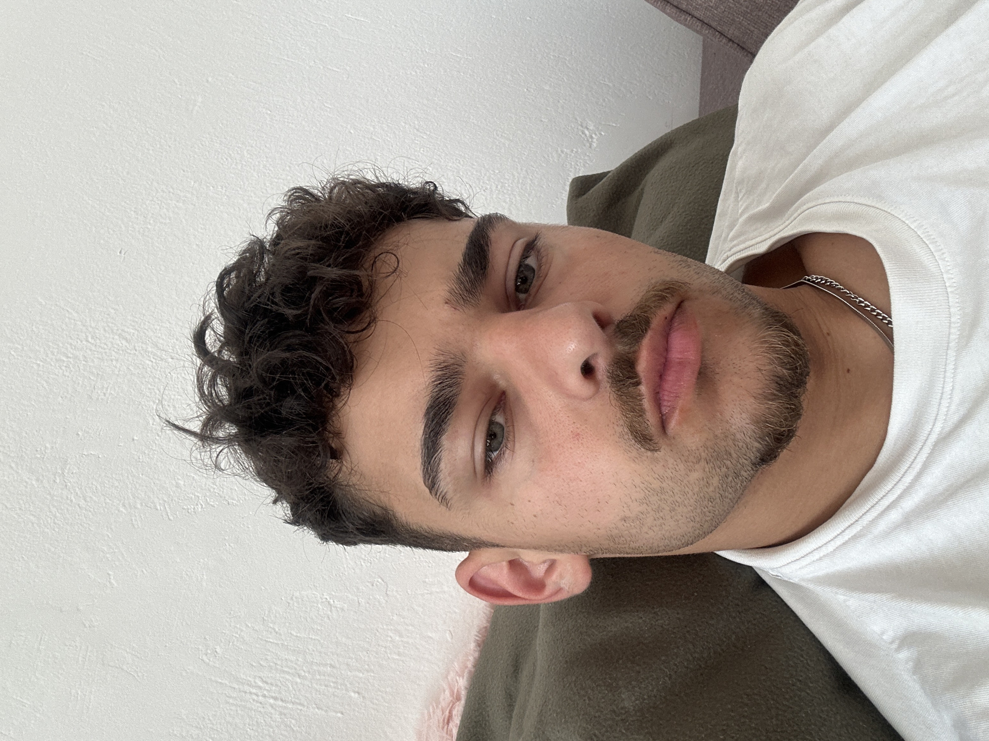 A close-up, angled portrait of a young man with dark, curly hair and a neatly trimmed beard and mustache. He is fair-skinned, has dark eyebrows, and appears to have greenish-gray eyes. He is wearing a white t-shirt and a silver chain necklace is visible at his neck. He is lying down on a soft, green surface (likely a couch or pillow). The background is a plain, white wall. There is no indication of the location being in Troyan, Bulgaria or any location.