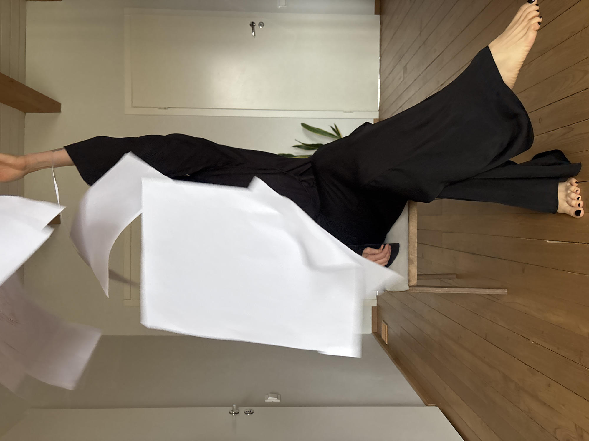 The image depicts an indoor scene, likely within a residence in Chaikovsky, Russia, capturing a person in a dynamic moment surrounded by falling papers. The primary subject is a person, whose face is not visible, shown from the mid-torso down. They are wearing loose, wide-leg black trousers. Their bare feet are visible, adorned with black nail polish; one foot is gracefully extended upwards, while the other rests on the light wooden floor. The person appears to be seated or reclining on a low piece of furniture, possibly a chair or bench, positioned on the right side of the frame.
The central activity involves numerous white sheets of paper that are scattered and appear to be in motion, either falling, being thrown, or floating through the air. Several of these papers exhibit motion blur, emphasizing their movement. One of the person's lower arms, visible on the left, is extended, suggesting interaction with or release of these papers.
The setting features a warm, light brown wooden floor with horizontal planks, covering the lower and right portions of the image. The walls are a plain, neutral light color, possibly off-white or beige. In the background, a white door with a silver handle is visible on the upper left, and a green-leafed plant adds a natural element to the minimalist interior. A wooden beam or shelf is also seen on the far left. The lighting is soft and diffused, characteristic of an indoor daytime scene. There is no visible text in the image.