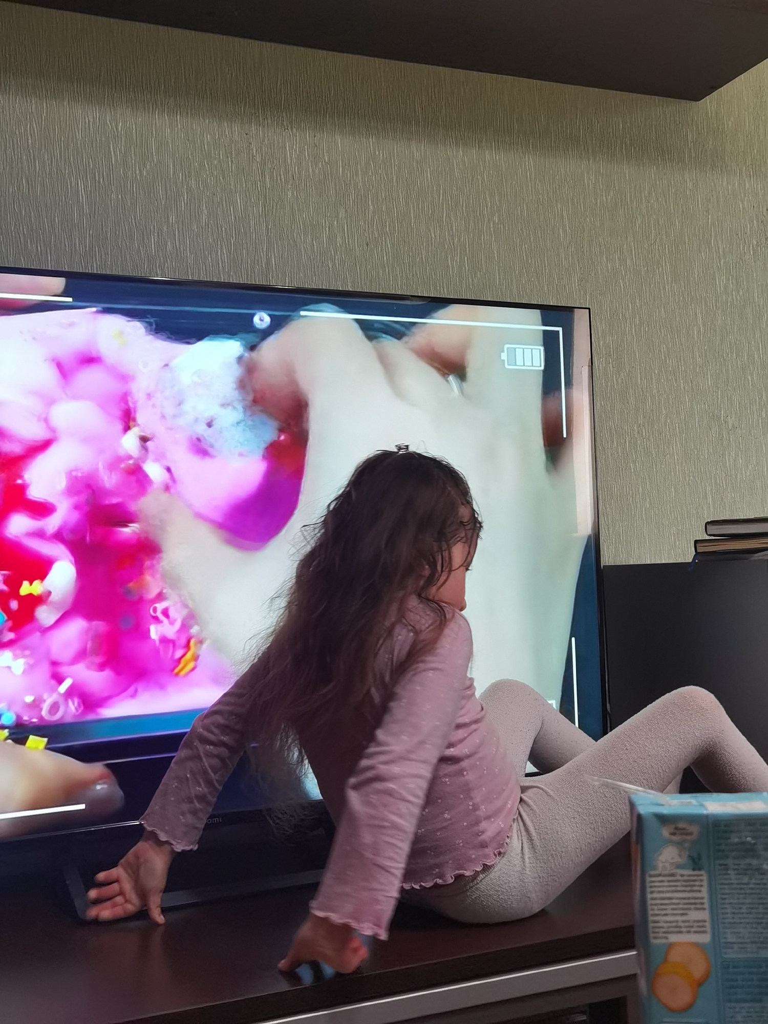 The image depicts an indoor domestic scene in the city of Chaikovsky, Russia, featuring a young child watching a large television screen. A child, appearing to be female with long, wavy brown hair, is seated on a dark brown media console or low table, facing the television with her back partially to the viewer and head turned towards the screen. She wears a light pink long-sleeved top with subtle white patterns and light grey textured leggings. Her right hand rests flat on the console surface near the television's stand, while her left hand is mostly obscured.
The central object is a large, flat-screen television displaying vibrant content. The left portion of the screen shows a chaotic arrangement of bright pink and red substances, possibly slime or liquid, intermixed with small, colorful, scattered objects, including yellow elements. The right portion of the screen features a lighter, creamy background where a hand is visible interacting with a foamy, blue-white substance and a pink object. A white rectangular frame is superimposed on the screen's content, with a fully charged battery icon (three bars) displayed in its upper right corner. White vertical and horizontal lines also mark sections of the display.
The television rests on a dark brown, polished media console. Above the television, a dark brown shelf is mounted on a wall covered in light brown/beige textured wallpaper with vertical striations. To the right of the television, a black monitor or electronic device is visible, topped with a stack of dark-spined books. In the lower right foreground, on the console, a light blue carton box is partially visible, featuring an image of two yellow-orange cookie-like items and text in an unidentified script. The overall lighting is soft and ambient, characteristic of an interior room.