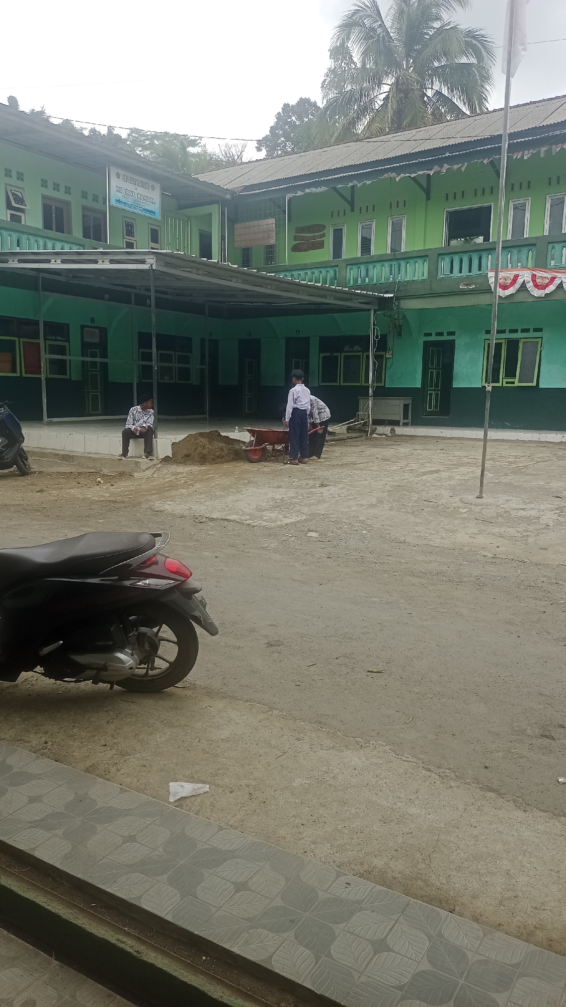 In Karyamandala, Indonesia, an outdoor courtyard of a green building, possibly a school or religious institution, is depicted. The building has a two-story structure with an overhang supported by pillars. The courtyard surface is paved with concrete and dirt. In the foreground, a dark-colored scooter is parked. Further into the courtyard, two individuals, one appearing to be a student in a white shirt and blue pants, and another in patterned attire, are near a pile of dirt and a red wheelbarrow. Another person in a patterned shirt and white cap is seated on a low wall. The sky is overcast, and palm trees are visible behind the building. The building displays signage with Indonesian text, including "SELAMAT DATANG" and "MTs. RAUDLATUL JANNAH". Decorative elements, such as banners and intricate green railings, are visible on the building's facade.
