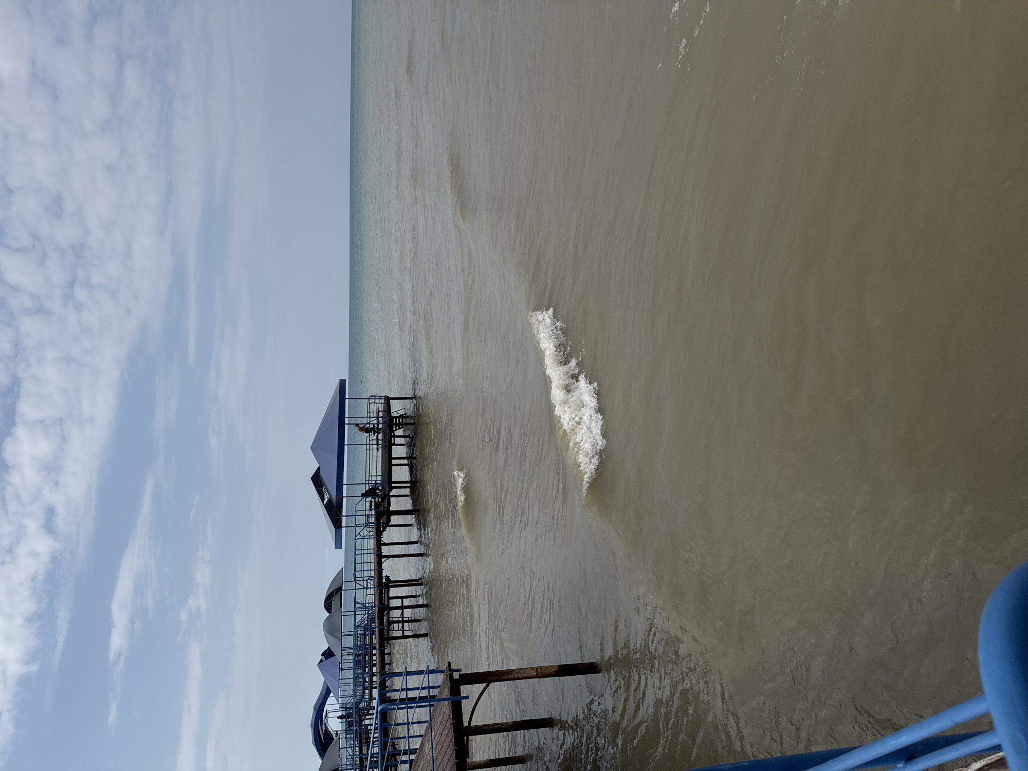 The image depicts a view from a wooden pier extending over murky water, likely a body of water near Sary-Oy, Kyrgyzstan. Several covered gazebos with blue roofs are visible along the pier, supported by wooden stilts. Two individuals are present on one of the gazebos, one appearing to be sitting while the other stands. Small waves are breaking near the pier. The sky above is cloudy, with a mix of white and gray clouds against a pale blue background. The overall impression is a calm, overcast day at a lakeside or seaside resort.