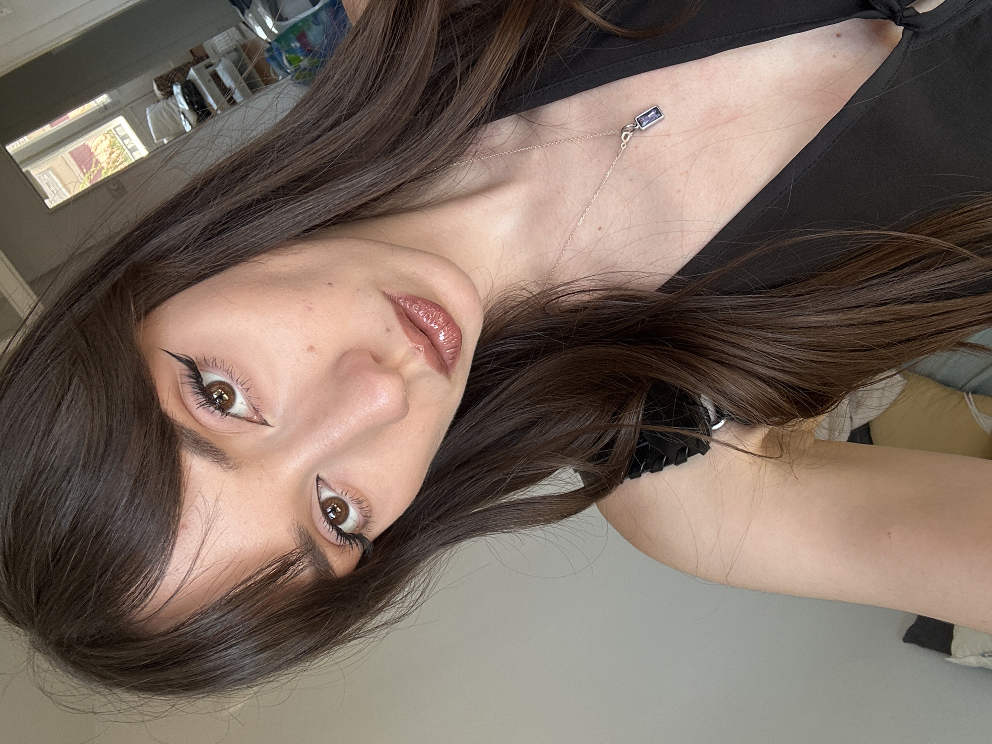 The image shows a close-up, selfie-style portrait of a young woman with long, dark brown hair and bangs. She is wearing a black top with thin straps and a pendant necklace with a rectangular purple stone. Her makeup includes winged eyeliner and shimmery, rosy-toned lipstick. The background is a plain, light-colored wall, and a blurred reflection of a room with windows and furniture is visible in a mirror on the right side of the frame. The lighting suggests it is daytime. The scene does not provide any specific visual cues to determine a location within the city of Belgrade, Serbia.