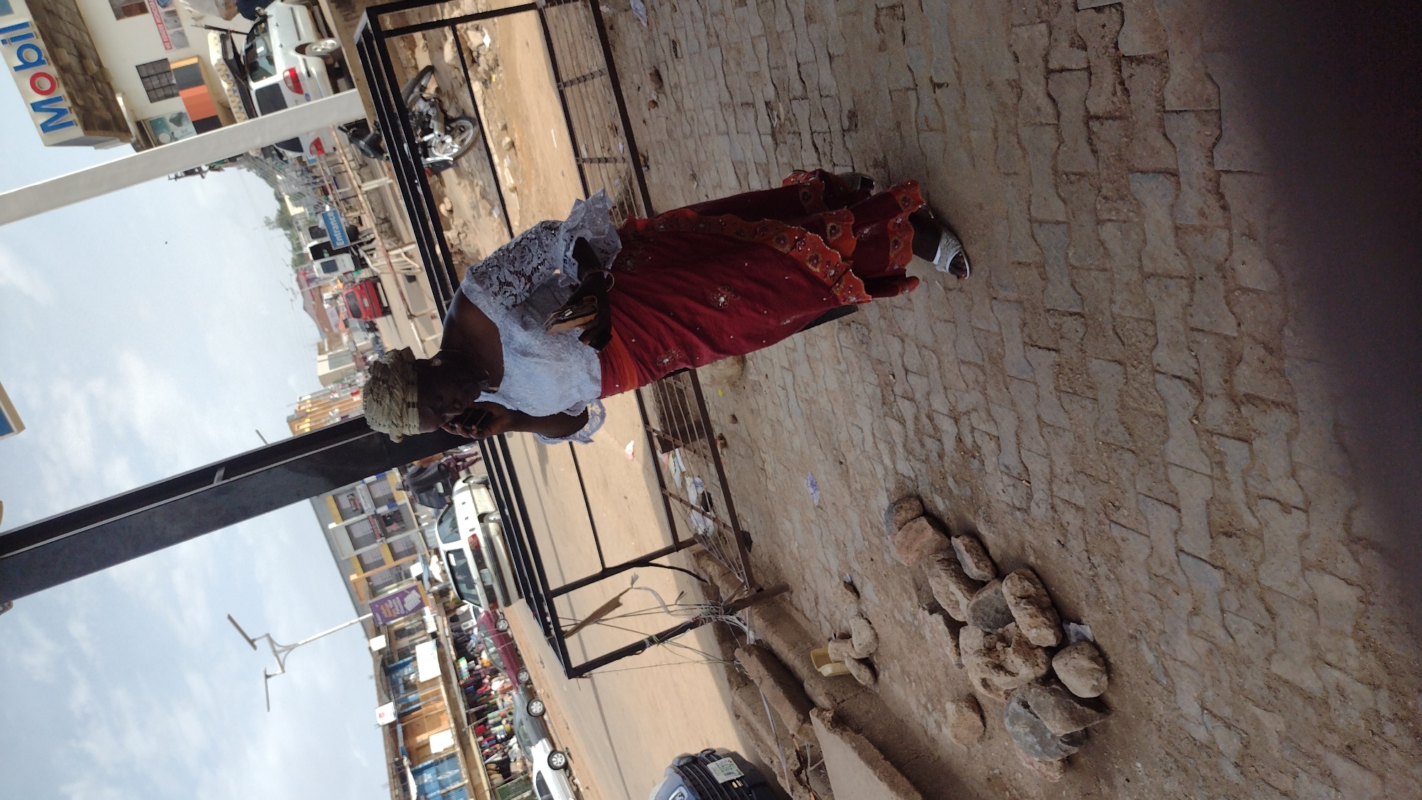 An elderly African woman is standing on a paved sidewalk, speaking on a mobile phone. She is wearing a white blouse with lace detailing, a red and orange patterned skirt, and a headwrap. She is located on a bustling street in Jos, Nigeria, evidenced by the "Mobil" gas station sign visible in the background. The sidewalk is lined with parked cars and open-front shops. Debris, including rocks, is scattered near the edge of the sidewalk. A metal frame structure is situated on the sidewalk nearby.