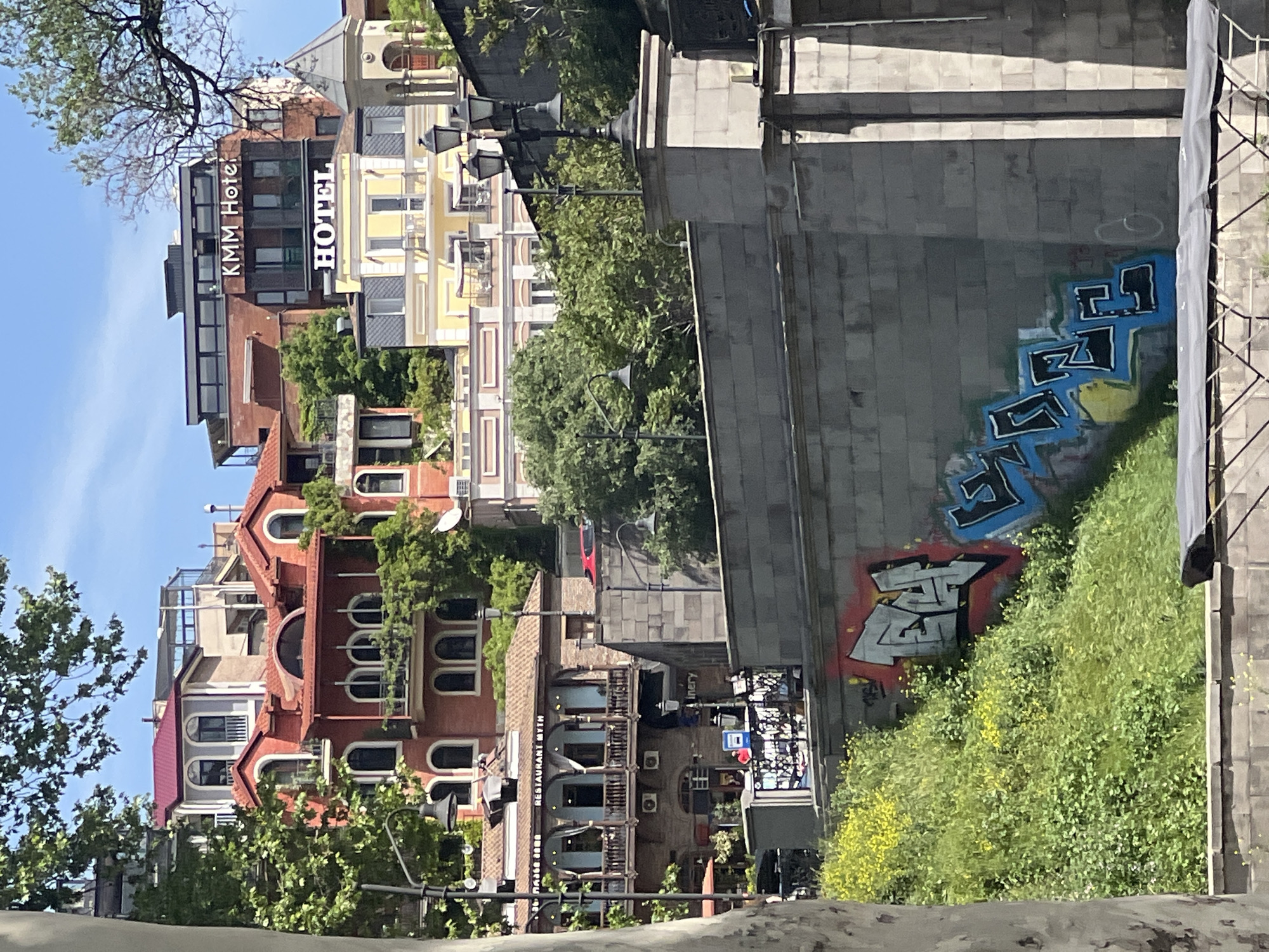 The image depicts a street scene in Tbilisi, Georgia, showcasing architectural diversity. Several buildings are visible, including a hotel with signage "KMM Hotel" and "HOTEL." The buildings vary in color, from red-brick structures to light yellow buildings. Some have multiple stories, arched windows, and balconies. Green foliage, including trees and vines, surrounds many of the buildings. A street lamp is positioned near a bridge-like structure made of grey stone, which is covered with graffiti. A hill covered in green foliage with yellow flowers is present in the scene. A portion of blue sky is visible.