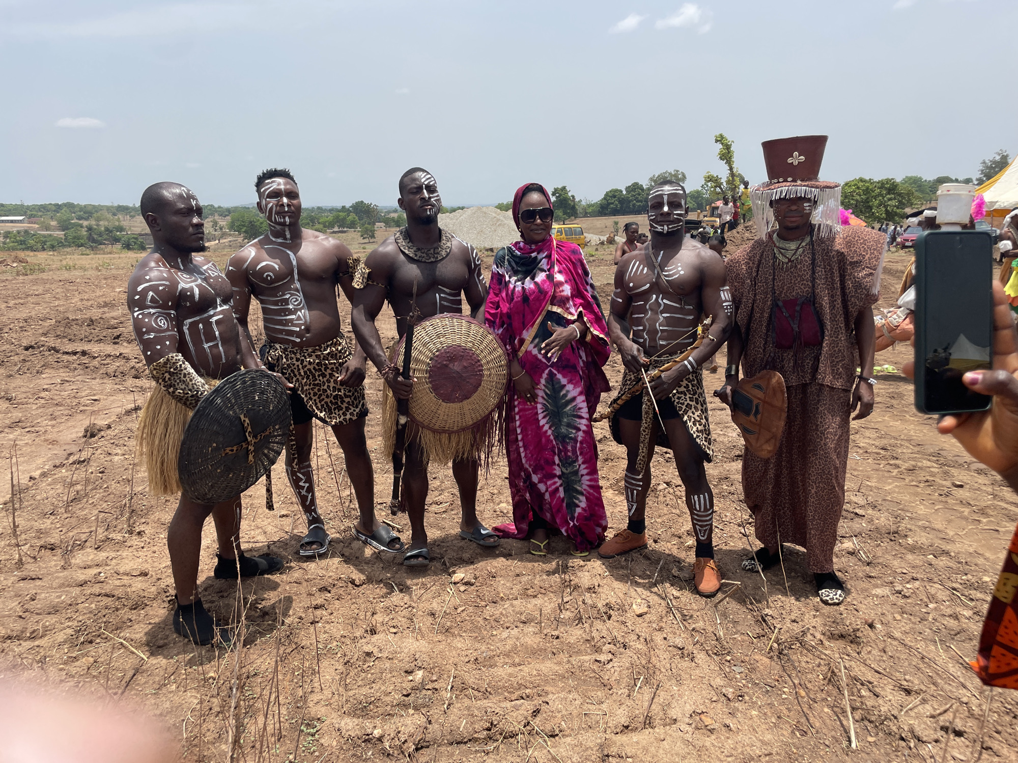 The image captures a group of six individuals standing in a dry, dusty field in Akwanga, Nigeria. Five of the people, all men, appear to be dressed in traditional attire that includes white body paint. Three wear leopard-print loincloths or wraps, and two have full outfits. They are holding items like shields and a fan. One woman, wearing a pink and black patterned robe, stands among them with sunglasses. Other people and temporary structures like tents can be seen in the background. A person holding a smartphone can be seen on the right. The landscape has dry grass and some trees in the distance under a partially cloudy sky.