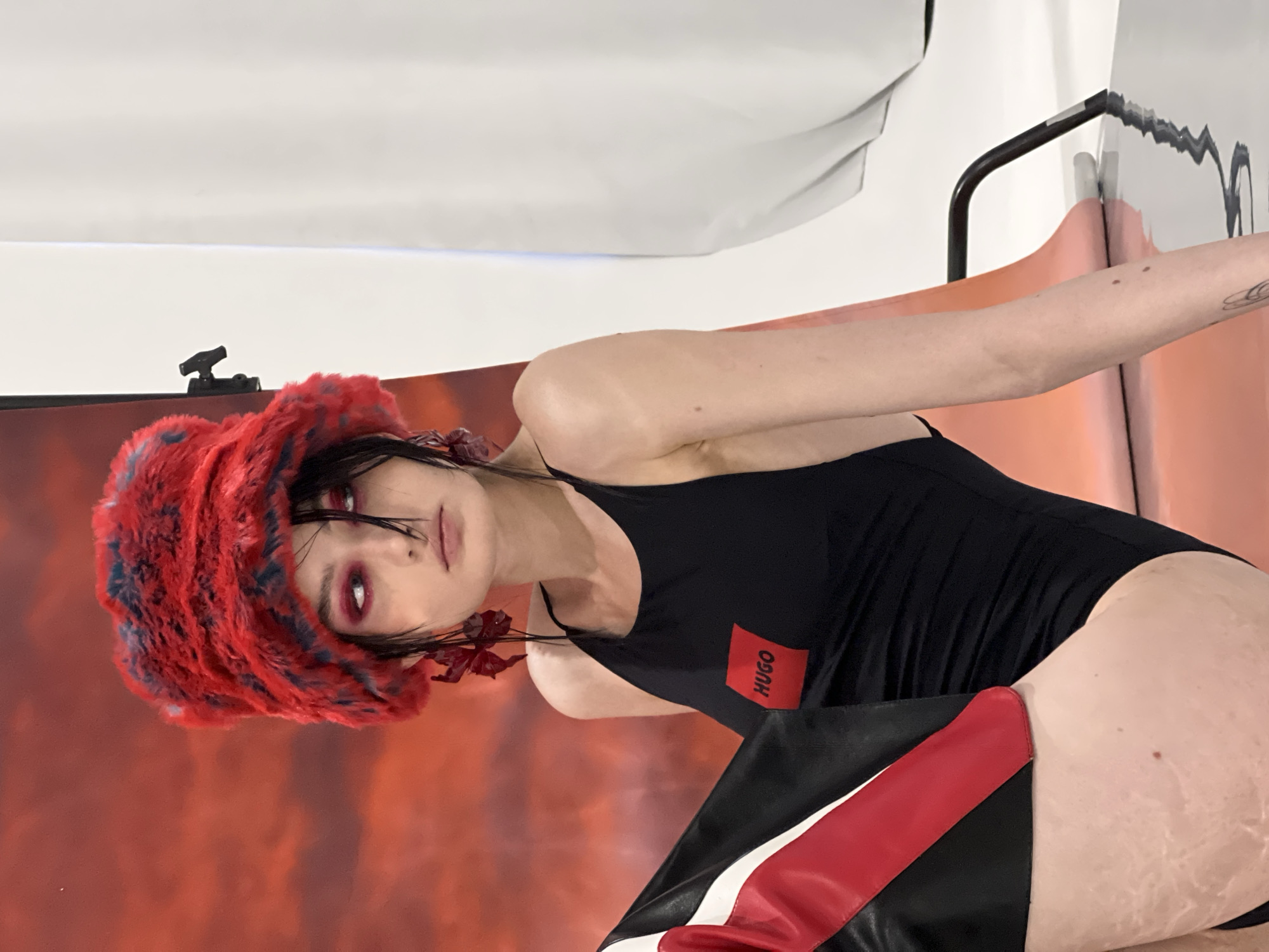 The image, presented with a 90-degree counter-clockwise rotation, depicts a person in an indoor studio setting, likely in Khimki, Russia, engaged in a fashion or artistic photoshoot. The subject has dark hair, striking red eye makeup, and dark lipstick, maintaining an intense and serious expression. They are wearing a fluffy red and blue hat and large red, floral-like earrings. The main garment is a black one-piece, possibly a swimsuit or bodysuit, featuring a prominent red rectangular patch on the chest with the white text "HUGO". The person's left arm is extended to the side, revealing a tattoo on the upper arm. Part of the lower body is visible, clad in a black garment with bold red and white accents, suggesting boots or chaps. The background is a vibrant, mottled red and orange, creating a dynamic backdrop that transitions into a plain white area at the top of the frame. Visible studio equipment, including a black clamp and other rigging, confirms the professional photography environment.