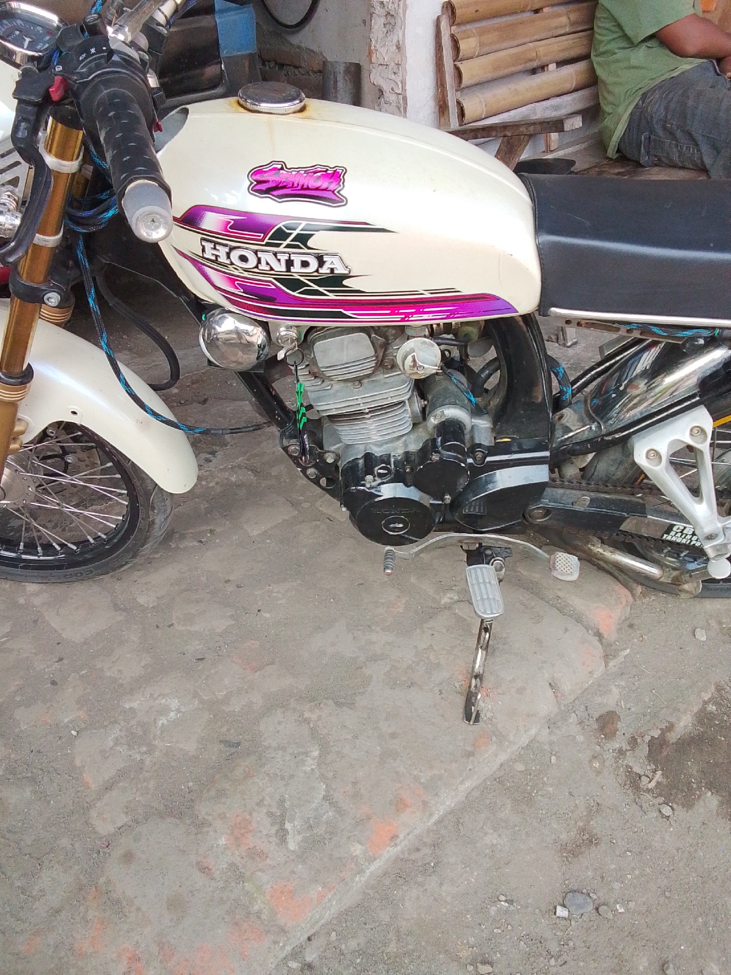 The image shows a close-up of a white Honda motorcycle parked on a concrete ground in Bulurejo, Indonesia. The motorcycle is the main subject, with its engine, fuel tank, front wheel, and handlebars clearly visible. The fuel tank features a graphic with the "HONDA" logo in white letters outlined in purple and pink. The motorcycle has gold-colored front forks and a black seat. In the background, part of a person wearing a green t-shirt and dark pants is seated on a bamboo bench. The environment appears to be outdoors, with a textured concrete ground and a brick wall in the background.