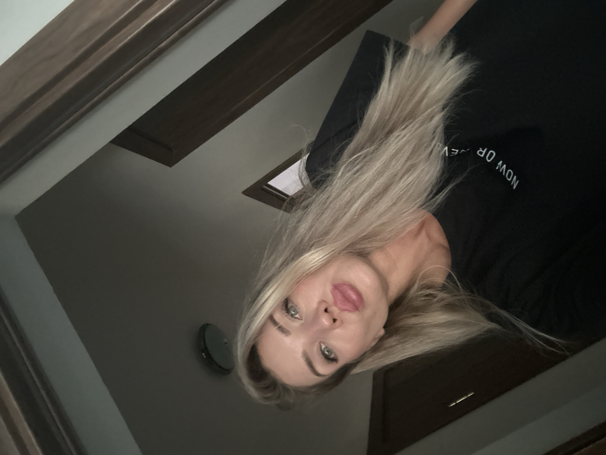 A selfie of a blonde woman is taken from a high angle, with her face positioned in the upper left quadrant of the frame. She is wearing a black t-shirt with white text that appears to read "NOW OR NEVER" in an arc. Her hair is long and blonde, and she is making a duck face. The background is dimly lit and appears to be an interior space with wooden trim and a ceiling light fixture. The setting is not identifiable as Armavir, Russia.