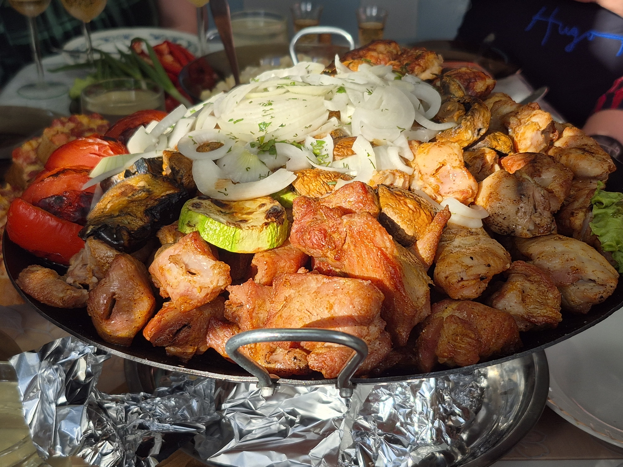 A close-up shot reveals a large platter of grilled meats and vegetables. On top, a generous pile of sliced onions, garnished with what appears to be chopped parsley, dominates the center. Surrounding the onions are pieces of grilled chicken and various vegetables, including red peppers, zucchini, eggplant, and tomatoes. The platter is supported by a stand covered in aluminum foil. In the background, there are wine glasses and other food items, indicating a social gathering. The presence of a table suggests the scene is indoors, possibly a restaurant. Based on the overall appearance, the photo captures a meal in a communal setting. It is not possible to confirm its origin from Votkinsk, Russia.