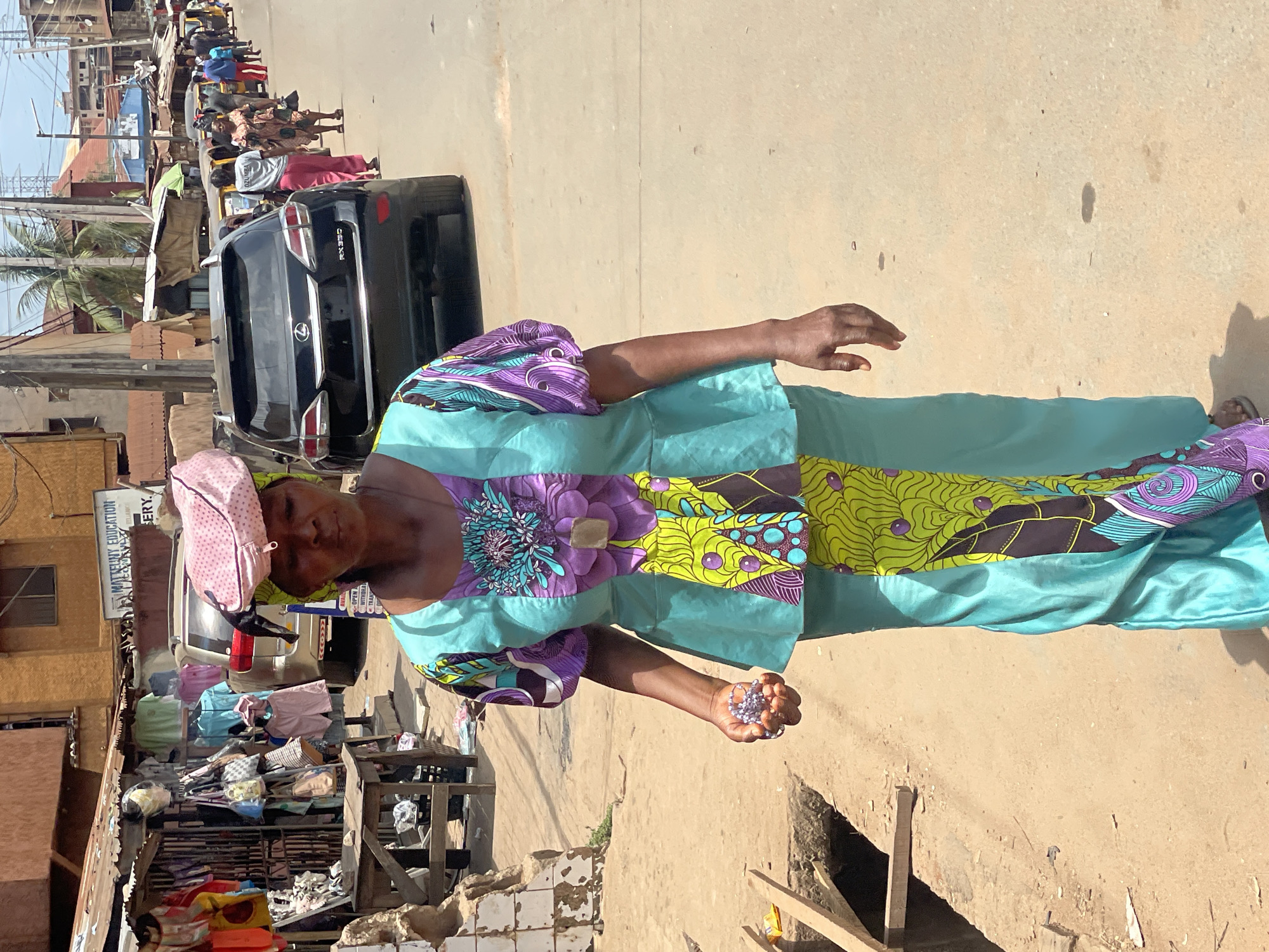 The image captures a vibrant street scene in Onitsha, Nigeria, featuring a woman in the foreground against a backdrop of a busy road and market activity under bright daylight.
In the foreground, a woman stands upright, looking directly at the camera with a neutral expression. She is dressed in a distinctive West African print outfit, comprising a long-sleeved top and skirt in teal, purple, and lime green patterns. A pink, textured head covering is tied on her head. Her right arm is extended slightly, while her left hand is held at waist height, grasping a small, shiny, possibly beaded item.
Behind her, a paved or dirt road extends into the distance, with several vehicles and people visible. A black Toyota RAV4 SUV is parked on the road, with its rear facing the viewer. Further down the street, a line of yellow commercial tricycles, commonly known as keke napep, are parked or moving slowly, occupied by multiple passengers. Buildings with corrugated iron roofs line both sides of the street, some appearing to be market stalls displaying various goods. Overhead, utility poles and electrical wires crisscross the sky.
Visible text includes "RAV4" on the back of the SUV and a partially obscured sign on a building that reads "MATERNITY EDUCATION NURSERY." Another sign shows fragments like "OPEN WITHDRAWAL TRANS...". The clear sky and strong shadows indicate a sunny day, likely mid-morning or early afternoon. The overall scene depicts typical urban life and commerce in a Nigerian city.