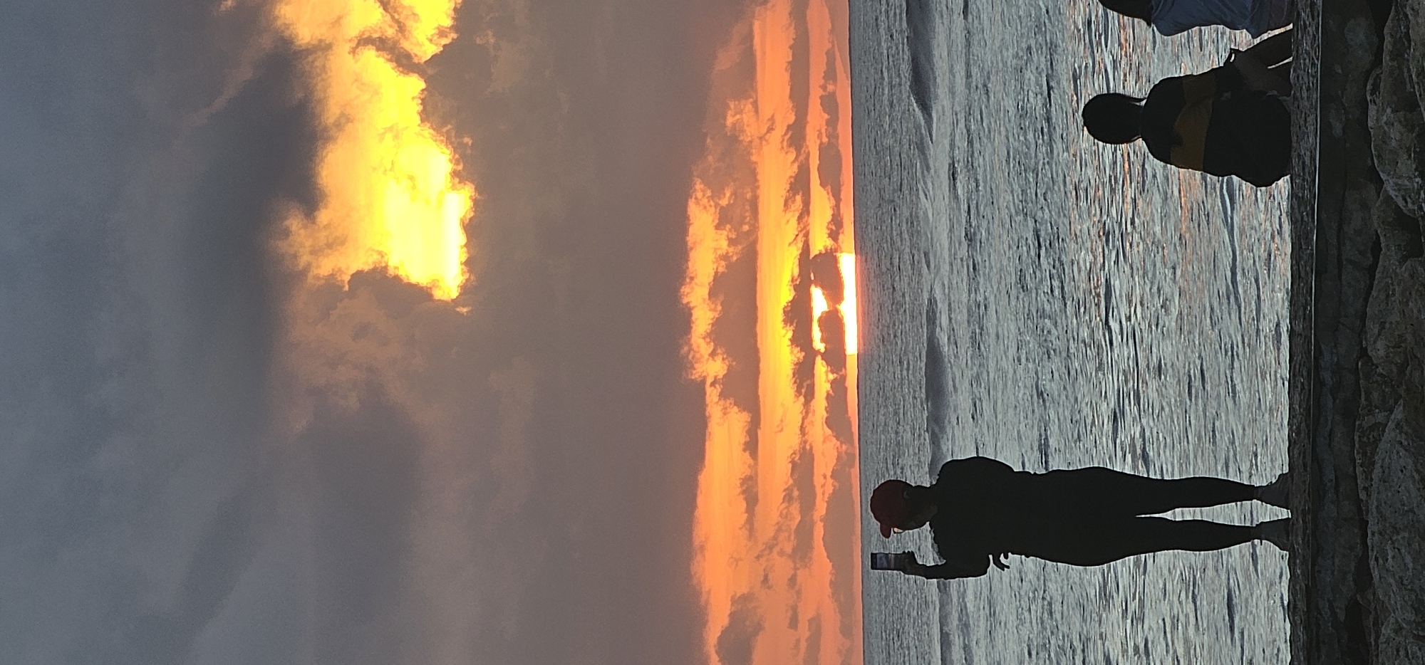 A vertically oriented, wide shot shows a sunset, ocean, and people. The sky is filled with clouds, with yellow light breaking through. To the right, the ocean reflects the orange and yellow hues of the sunset. Along the right edge of the frame, two figures stand on a rocky structure. The person closest to the camera is silhouetted, appearing to be taking a photo with a mobile phone. A second person is sitting nearby. The image was taken in Denpasar, Indonesia.