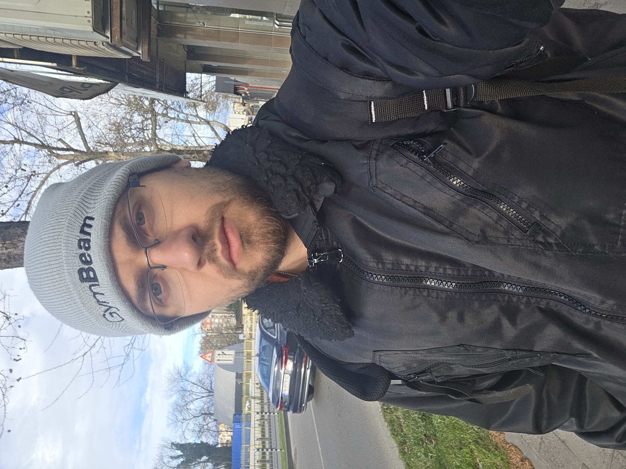 The image shows a person in Plovdiv, Bulgaria, wearing a grey beanie with the word "GymBeam" on it, glasses, and a black jacket. The person is outdoors, standing on a sidewalk. A black car is visible on the road behind the person, along with a road sign. Trees are visible in the background, some without leaves. The sky is partially cloudy.