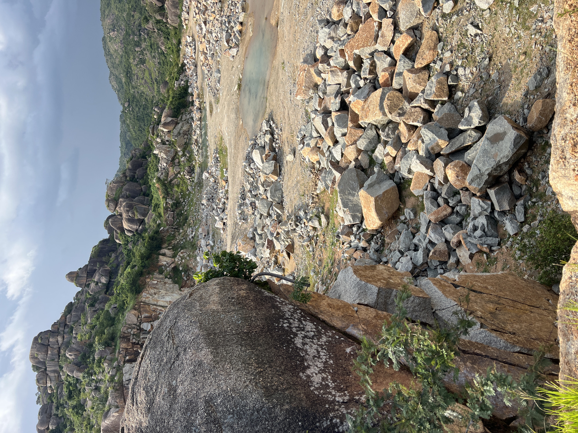 The image depicts a rocky, hilly landscape with scattered boulders and vegetation. In the foreground, a large, dark boulder with lichen growth is prominent. The terrain is uneven, with piles of smaller rocks and a dirt path winding through. There are small pools of water visible in depressions on the ground. In the background, more hills covered in rocks and greenery are visible, with a cloudy sky overhead. The scene appears to be a natural, undeveloped area. This type of terrain is common in and around the city of Jos, Nigeria, particularly in areas known for their granite rock formations and mining activities. Specifically, the landscape resembles areas like the Shere Hills or other quarry sites surrounding Jos. There are no people, animals, or man-made structures visible in the image.