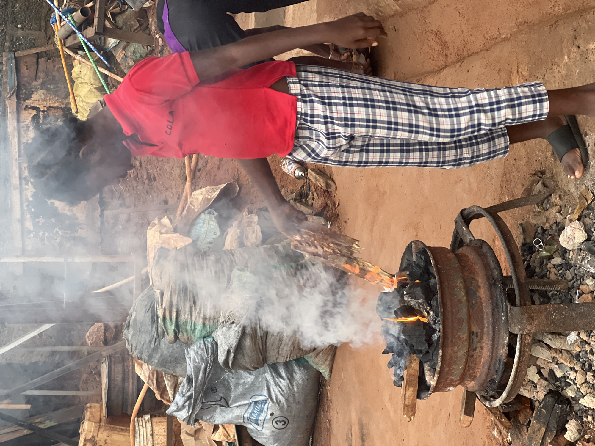 In Nkpor, Nigeria, a young individual in a red polo shirt with "C.O.L.A" printed on it and blue and white checkered trousers stands next to a makeshift charcoal grill. The grill, constructed from a rusty metal rim set on a stand, is filled with burning charcoal and emits a significant amount of smoke. The individual is holding a piece of burning wood over the coals, possibly to start the fire or for cooking. In the background, several large sacks, some appearing to contain materials, are piled up. The ground is made of reddish-brown earth, typical of the region. The overall scene depicts an outdoor, somewhat rustic setting where charcoal is being used for fuel.