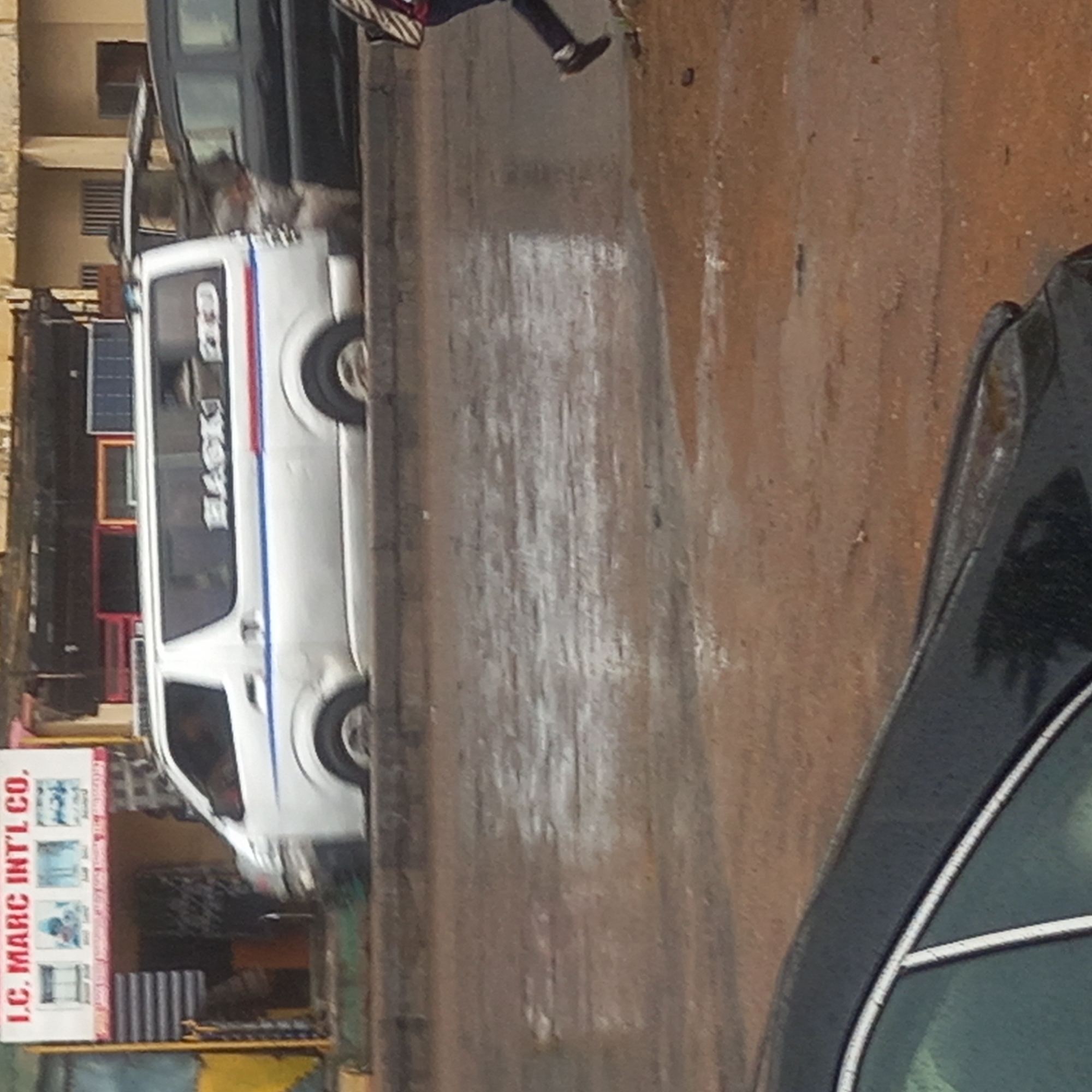 The image captures a street scene, likely during the daytime, with a wet ground surface suggesting recent rain or damp conditions. The setting appears to be a commercial or residential area.
In the mid-ground, a white passenger van, possibly a public transport minibus, is a prominent subject. It features distinctive blue and red stripes running horizontally along its side. On its side window, the word "BLACK" is visible in black text with a red outline, and "RED" is written in smaller red script above it, indicating a potential route or company name ("BLACK RED"). The van's wheels are visible, and it appears to be parked or moving slowly.
To the far right, the front passenger side of a black car, including part of its window and roofline, is partially visible.
A person is seen in the upper center of the image, walking away from the camera. They are wearing dark trousers and dark shoes.
On the left side of the street, a building is visible, and near its entrance, several rectangular objects resembling solar panels are displayed. Below these, a white sign stands vertically, bearing the text "I.C. MARC INT'L CO." in red letters. Smaller, less discernible text and images are also present on the sign. The overall scene depicts typical daily activity on a street.