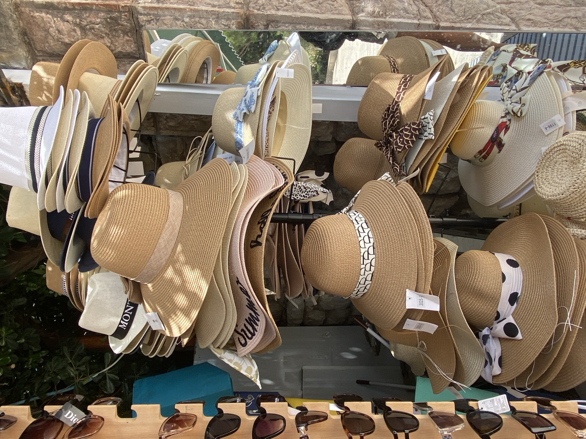 In Petrovac centar, Montenegro, a stall displays a variety of straw hats and sunglasses. The hats are stacked in multiple tiers, with the majority being wide-brimmed and made of woven straw. Several hats are adorned with ribbons in various patterns, including polka dots, abstract designs, and floral prints. Some hats also feature lettering, such as "MONTE" on a white fedora.
To the left of the hats, a wooden rack holds several pairs of sunglasses. These sunglasses have dark lenses and frames in shades of brown and black. The background of the scene consists of a stone wall, lush green foliage, and hints of a building's exterior. Sunlight casts shadows on the hats, indicating an outdoor setting. The overall impression is a retail display, likely in a tourist-oriented area.