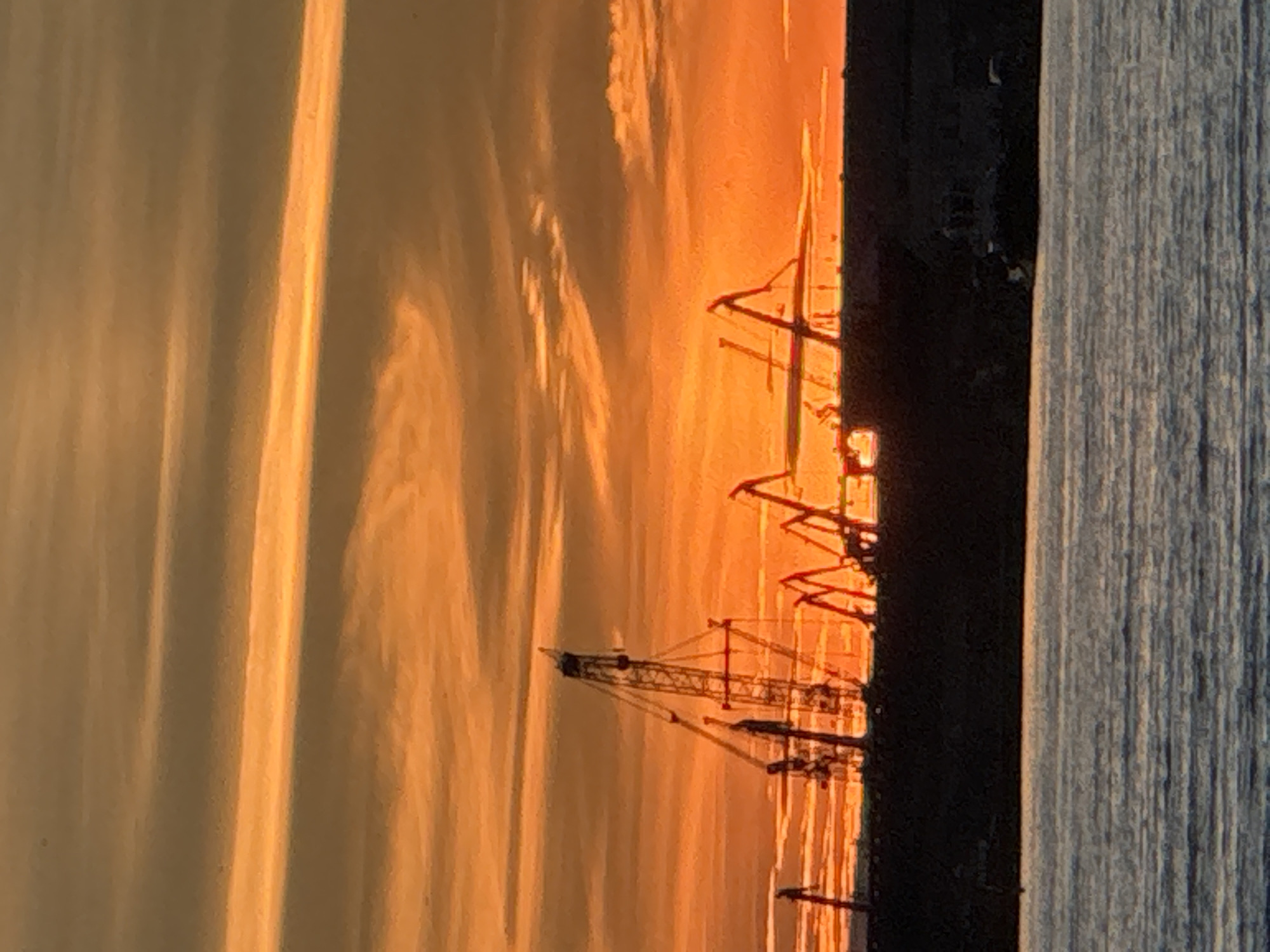 The image shows a harbor scene at sunset in Tallinn, Estonia. The sky is a vibrant orange and yellow, with horizontal clouds streaking across it. In the foreground, dark silhouettes of cranes and industrial structures are visible against the bright sky. The water in front of the harbor is dark and reflective. There are no people or vehicles clearly visible in the image. The overall impression is one of industrial activity juxtaposed with the natural beauty of a sunset.