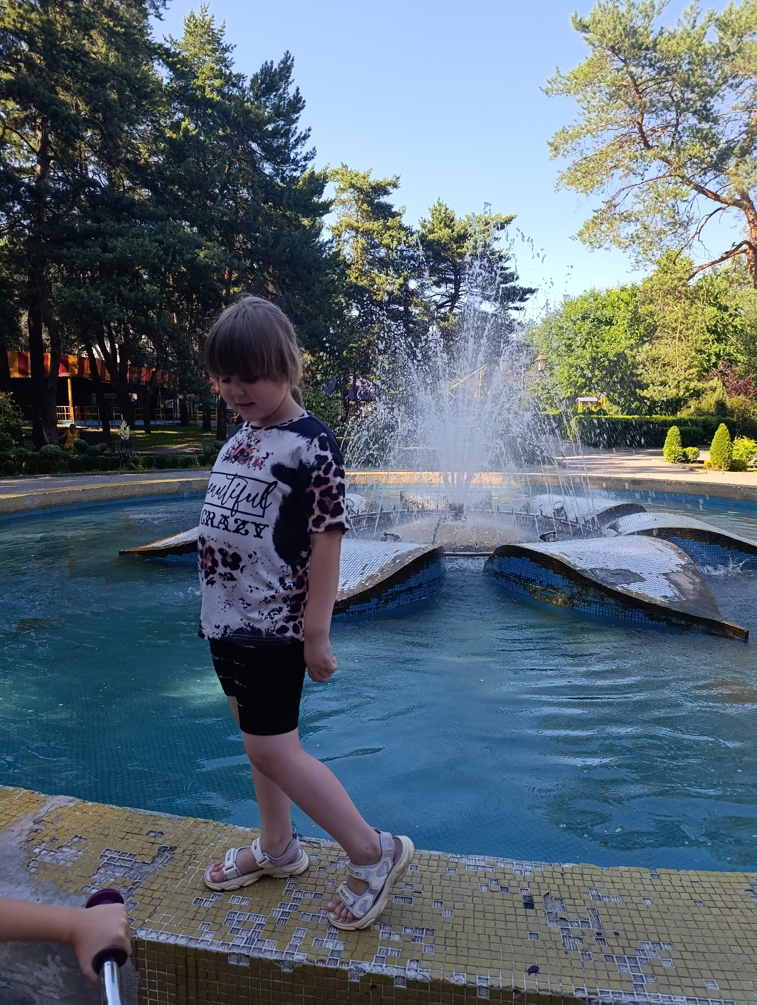 A young child, with light brown hair and bangs, stands on the edge of a tiled fountain in what appears to be a park or garden. The child is wearing a black and white t-shirt with a leopard print and text that reads "beautiful crazy," black shorts, and light-colored sandals. The child is looking down towards the water, with a neutral expression. The fountain itself is a large, circular structure with water spraying upwards from its center. The water in the fountain is a vibrant blue, and the fountain basin has decorative, petal-like structures submerged in the water, covered in blue and white tiles.
The setting is outdoors, with lush green trees visible in the background. The sky is clear and blue, suggesting it is a sunny day, likely late morning or afternoon given the lighting. The ground around the fountain is paved with yellow tiles. In the foreground, a hand and part of a scooter can be seen, hinting at another person or activity just out of frame. The overall atmosphere seems to be one of casual recreation and enjoyment of a pleasant day.