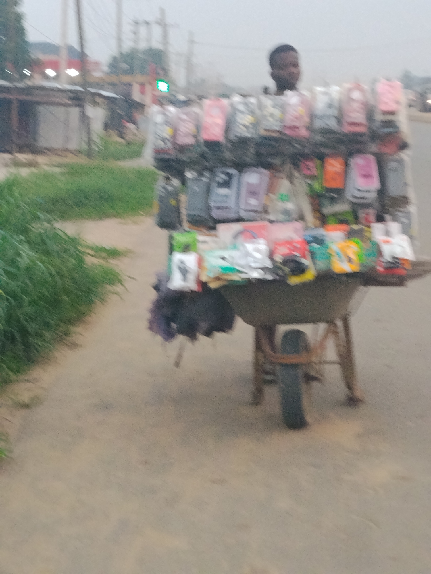 The image depicts a street scene in Ura Nta Umuarandu, Nigeria. A young man stands behind a wheelbarrow converted into a mobile phone accessory stand. The stand displays a variety of phone cases and accessories, packaged in plastic. The wheelbarrow is positioned on the side of a road with a green grassy area to the left. In the background, buildings and utility poles are visible under a hazy sky. A traffic light is also visible in the background showing a green light.