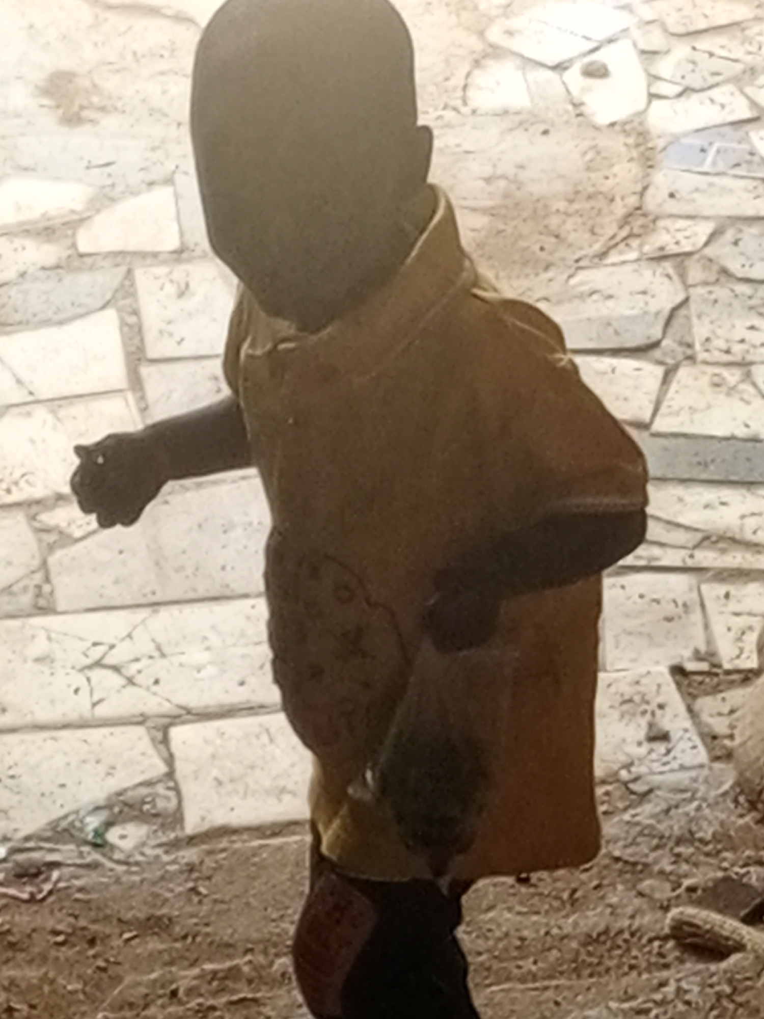 A child wearing a yellow shirt and dark pants stands on a dirt and stone ground in Bukuru, Nigeria. The child's face is obscured by shadow. The child's right arm is extended, and their hand is clenched into a fist. The background consists of a tiled wall and scattered debris.