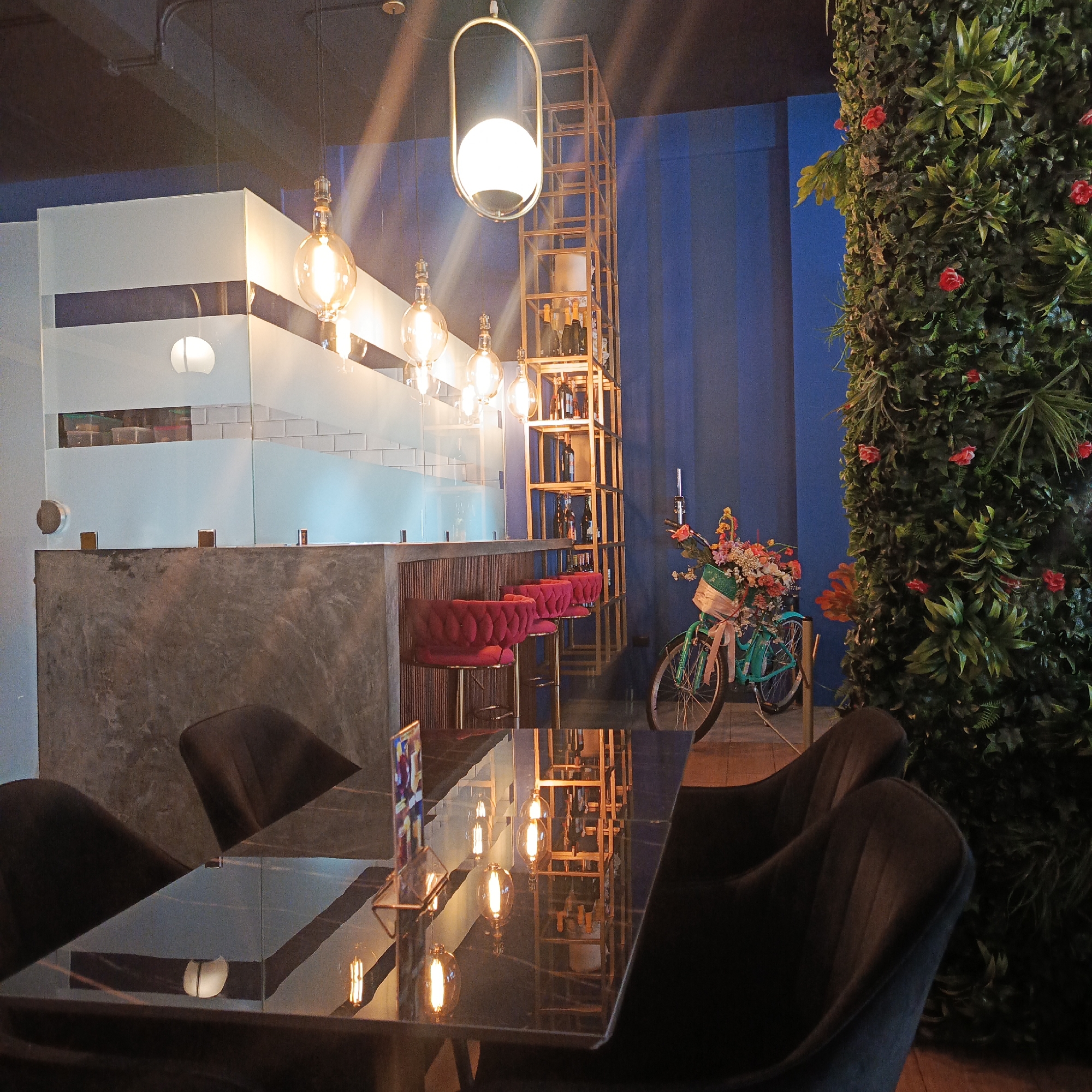 The image shows the interior of a cafe in Caracas, Venezuela. The cafe has a modern and chic design, with a bar area, seating, and a decorative wall of plants.
In the foreground, a reflective glass-top table is set with dark chairs around it. The table surface reflects the ambient lighting, creating a shimmering effect.
Behind the table, a polished concrete bar with a wooden front is visible. Several red velvet bar stools with gold legs are lined up against the bar, suggesting a seating area for patrons.
Further back, a tall, gold-colored shelving unit displays bottles, possibly wine or spirits, and some decorative items. The wall behind the shelving unit is painted a deep blue.
To the right, a striking vertical wall covered in lush green foliage and small red flowers creates a natural, vibrant backdrop. A vintage turquoise bicycle adorned with a basket full of colorful flowers is positioned in front of the plant wall, adding a whimsical touch to the decor.
The lighting in the cafe appears to be a mix of warm, vintage-style Edison bulbs hanging from the ceiling and a larger, modern pendant light. The overall atmosphere is dim and intimate, with a focus on stylish design elements and a blend of modern and natural aesthetics. No people are visible in the image.