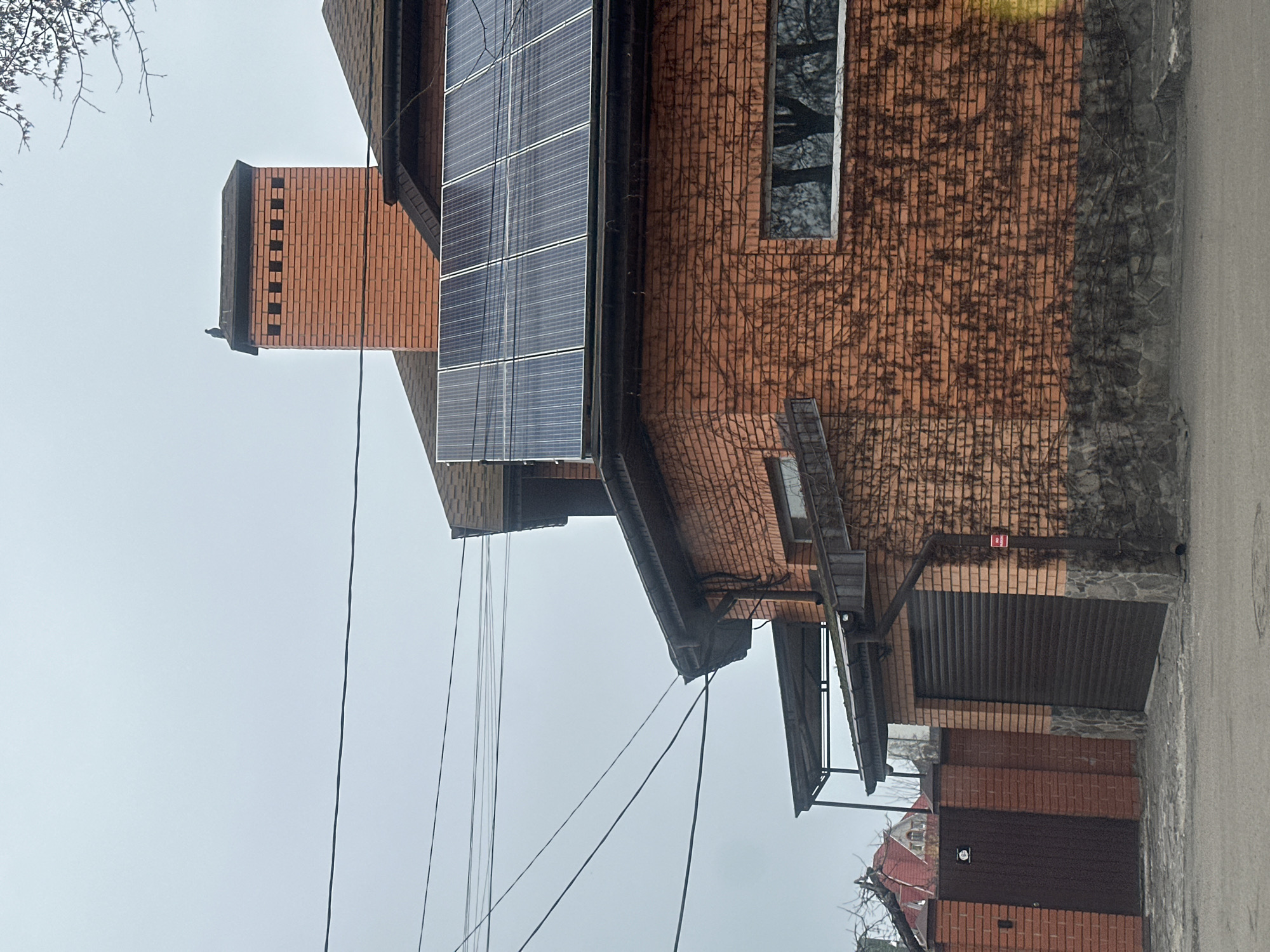 The image shows a building in Kyiv, Ukraine, constructed primarily of reddish-brown brick. The building features a chimney and a section of solar panels on the roof. Windows are visible in the brick facade. The wall of the building has patches of vines. A metal garage door is attached to the building. Power lines are visible in the sky. The sky appears overcast, suggesting a cloudy day.