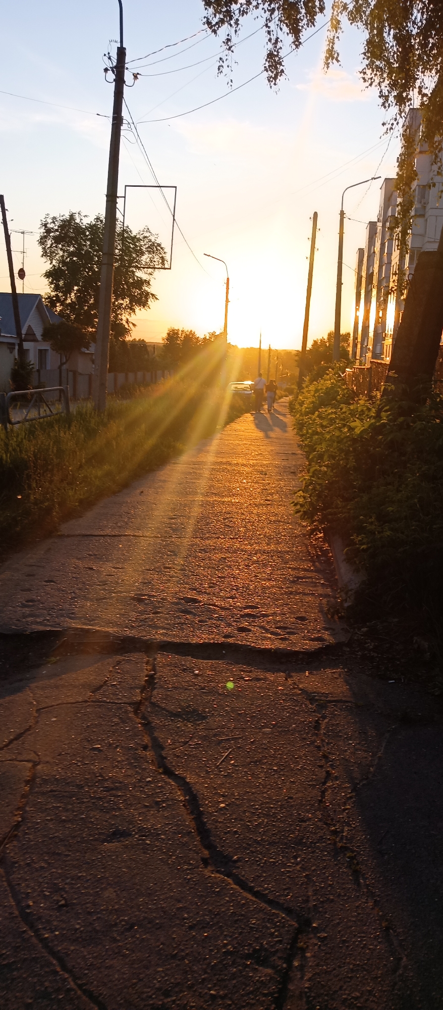 The image depicts a ground-level perspective of a cracked asphalt or concrete pathway at sunset, within the city of Chaikovsky, Russia. The sun, located low on the horizon in the upper-middle, emits intense golden-orange light, producing prominent lens flares that radiate across the scene and cast long shadows forward from subjects. The sky in the upper third transitions from light blue at the zenith to bright yellow and orange at the horizon. Overhead, multiple black power lines are visible, extending between utility poles. Hanging branches with small, dark leaves from a deciduous tree are present in the upper right. On the left side of the pathway, a prominent dark utility pole stands vertically, equipped with several wires and a horizontally oriented, empty, rectangular metal frame attached below mid-height. Further left and slightly in the background, another utility pole with an antenna-like structure and a red circular object (possibly a sign) is partially visible. Behind these poles, a fence lines the path, and a residential house with a blue-grey gabled roof and light-colored walls is visible. Beyond the house, additional trees and distant structures are present. Green grass and low-lying vegetation grow along the immediate left edge of the pathway. On the right side of the pathway, a series of dark utility poles extends into the distance. Adjacent to these poles are multi-story buildings, appearing light-colored, with numerous windows reflecting the direct sunlight. Dense green foliage, including bushes and a thick tree trunk, borders the immediate right edge of the path, creating areas of deep shadow. The pathway itself occupies the majority of the lower two-thirds of the image. It is visibly worn, characterized by numerous cracks, undulations, and small indentations, indicative of aging and use. The surface is illuminated by a strong golden-orange hue from the setting sun, highlighting its texture. A small, bright green circular artifact (lens flare) is visible on the path in the lower-middle-right quadrant. In the midground, two individuals are walking away from the camera, along the pathway towards the sunset. They appear as silhouettes or are backlit by the intense sun. One individual is taller, consistent with an adult male, and the other is shorter, consistent with an adult female. Their elongated shadows stretch onto the path ahead of them. A light-colored sedan is parked on the left side of the path, positioned between the sun's rays and the walking individuals. The scene conveys a