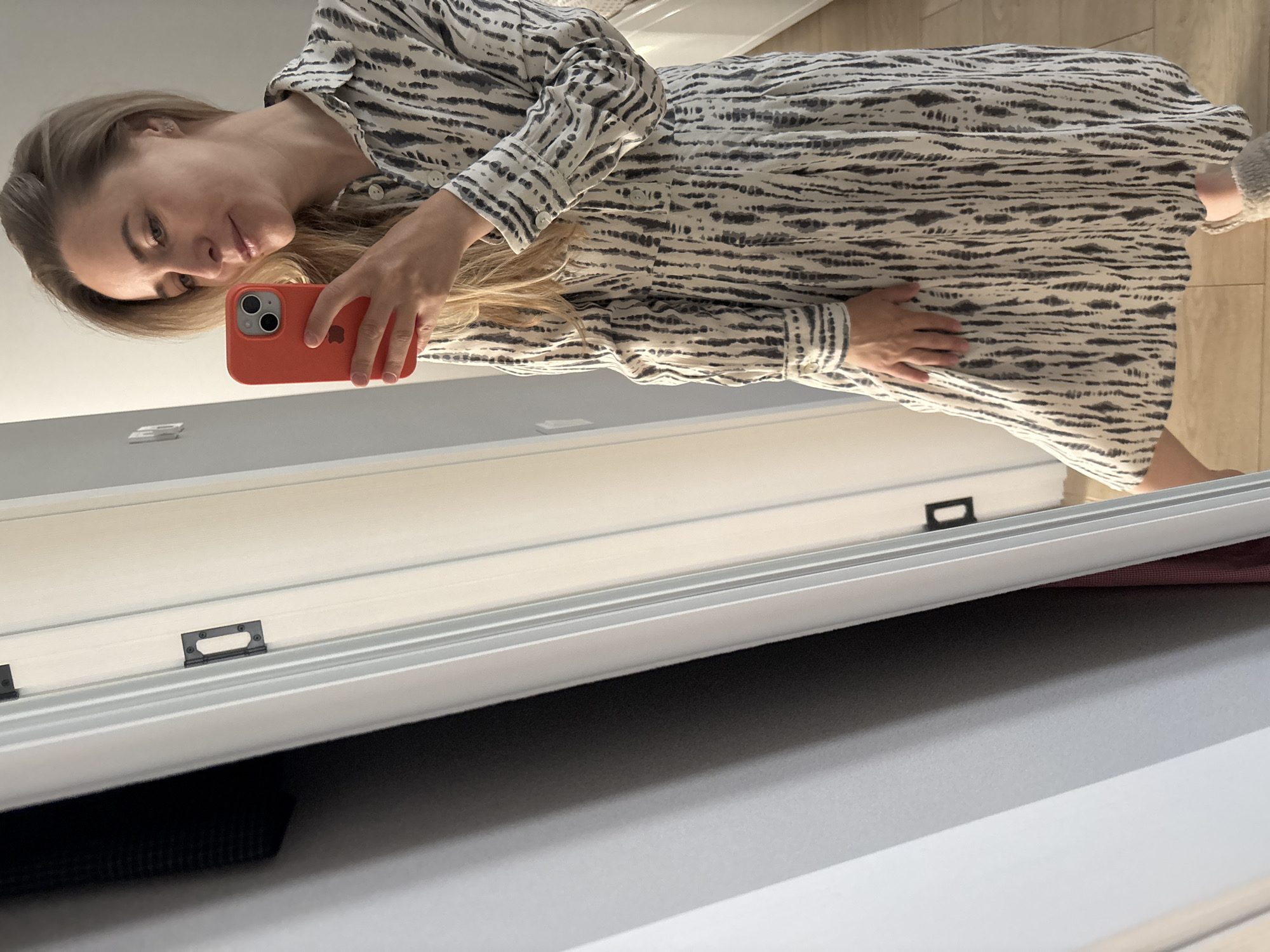 A woman with blonde hair is taking a mirror selfie with a red iPhone. She is wearing a long-sleeved dress with a black and white pattern. The background appears to be an indoor setting with a light-colored wall. There is no indication of the city Chaikovsky, Russia.