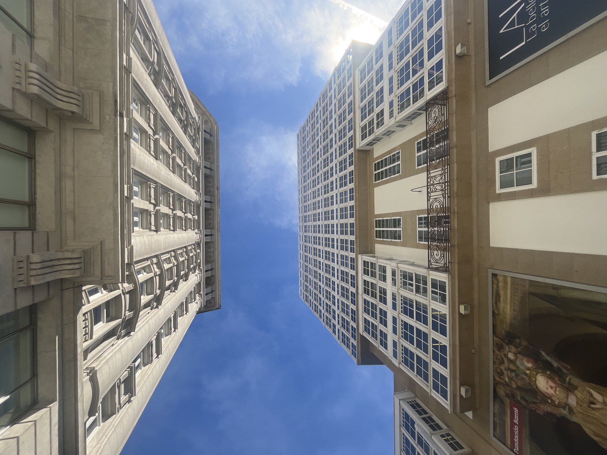 The image shows a low-angle shot of the Maria Pita Square in A Coruña, Spain, featuring the imposing architecture of the City Hall building. The square is famous for its iconic circular design and the statue of María Pita at its center, although these are not directly visible in this particular frame. Instead, the focus is on the facades of the surrounding buildings, which are characterized by their traditional Galician style with large glassed-in balconies, known as "galerías." The sky above is a vibrant blue with scattered white clouds. One of the buildings displays a large mural art piece on its lower level, and a sign partially visible reads "Fundación Barrie." The architecture is grand and ornate, reflecting the historical significance of the location.