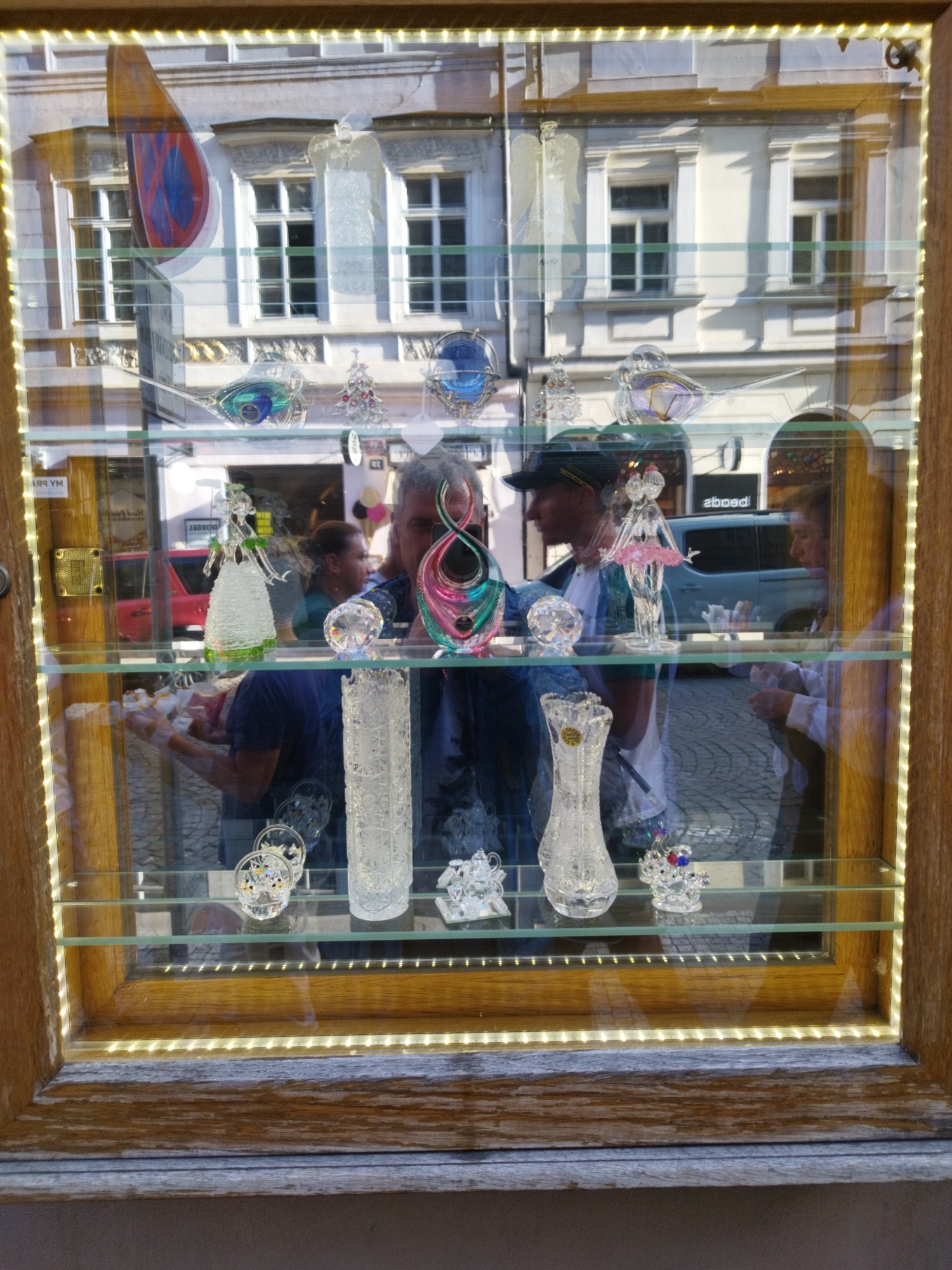 The image is a medium shot of a glass display case in a shop window, likely in Prague, Czechia. The display case has a wooden frame and is illuminated by warm LED lights around the edges. Inside, several shelves hold various glass figurines and decorative items.
On the top shelf, a large, colorful abstract glass sculpture is visible on the left, reflecting the building across the street. To its right, a clear glass angel figurine is displayed, with a small, sparkling Christmas tree next to it. Further to the right, a blue glass object, possibly a bird or orb, is present.
The middle shelf features a clear glass figurine of a person in a dress on the left. In the center, a man is reflected in the glass, wearing a black baseball cap and a blue t-shirt. He appears to be taking a photo of the display. To his right, a pink and clear glass figurine of a dancer is displayed.
The bottom shelf contains a tall, clear glass vase with intricate patterns. To its left and right are smaller glass decorative pieces, some of which appear to be flowers or animals. Reflections of people and the street scene outside are visible throughout the display. A red car is reflected in the glass on the far left, and a light-colored vehicle is visible in the background on the right. The ground outside appears to be cobblestone.