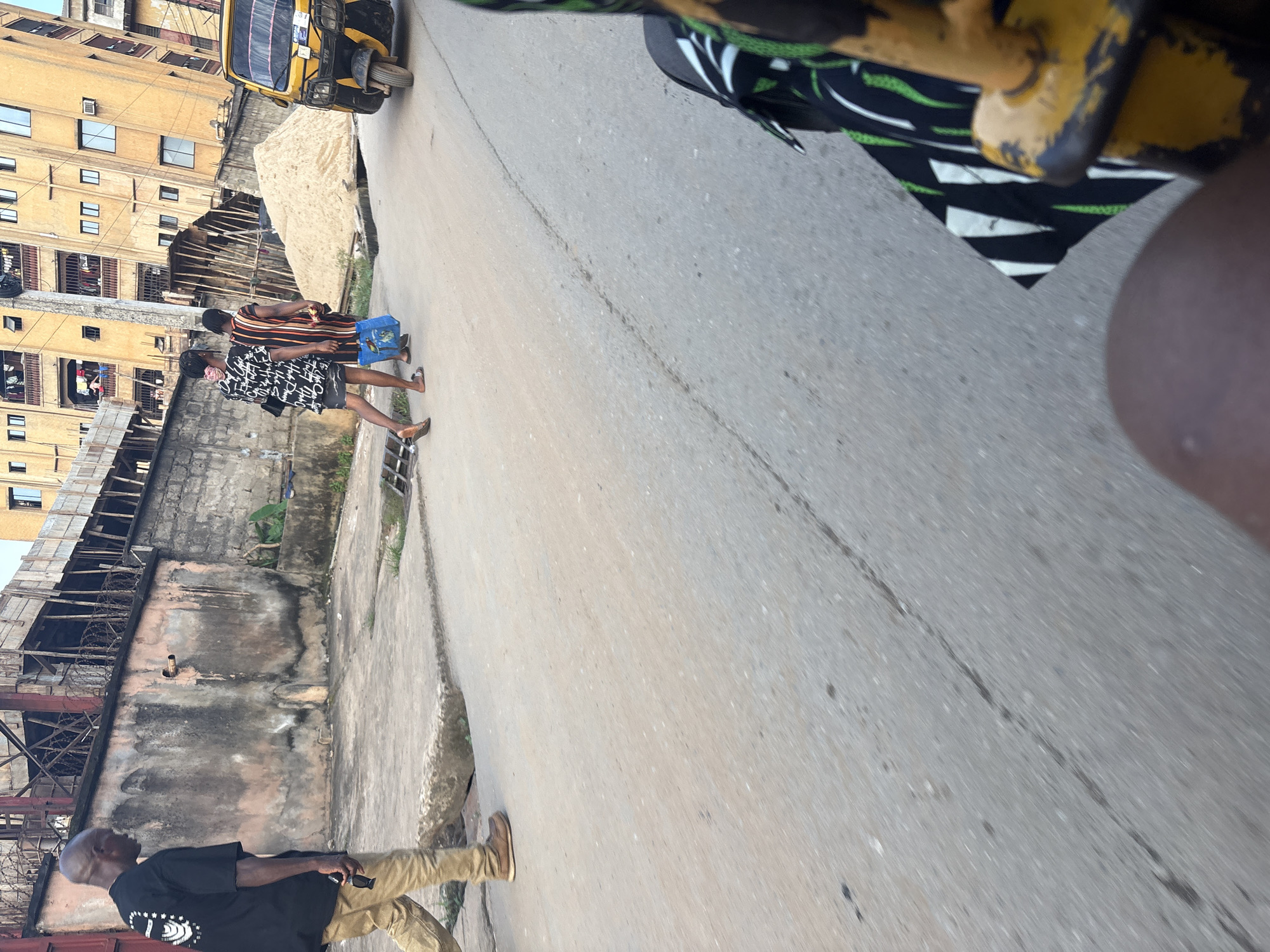 The image captures a street scene in Onitsha, Nigeria, with a focus on pedestrian movement and the urban environment. In the foreground, a person's leg and the back of a yellow three-wheeled vehicle, likely a commercial taxi, are visible, suggesting the photo is taken from within or immediately behind such a vehicle. On the right side of the street, a large pile of sand is situated next to a building under construction. Further down the street, two young women walk away from the viewer, one in a black and white patterned dress and the other in a striped dress, with the woman in the patterned dress carrying a blue bag. To their left, a man in a black t-shirt and khaki pants walks towards the viewer, holding sunglasses in his hand. The background features multi-story residential buildings with balconies and exposed structures, indicative of ongoing development or renovation. The street itself is paved with asphalt, showing signs of wear and tear. The overall atmosphere suggests a busy urban setting.