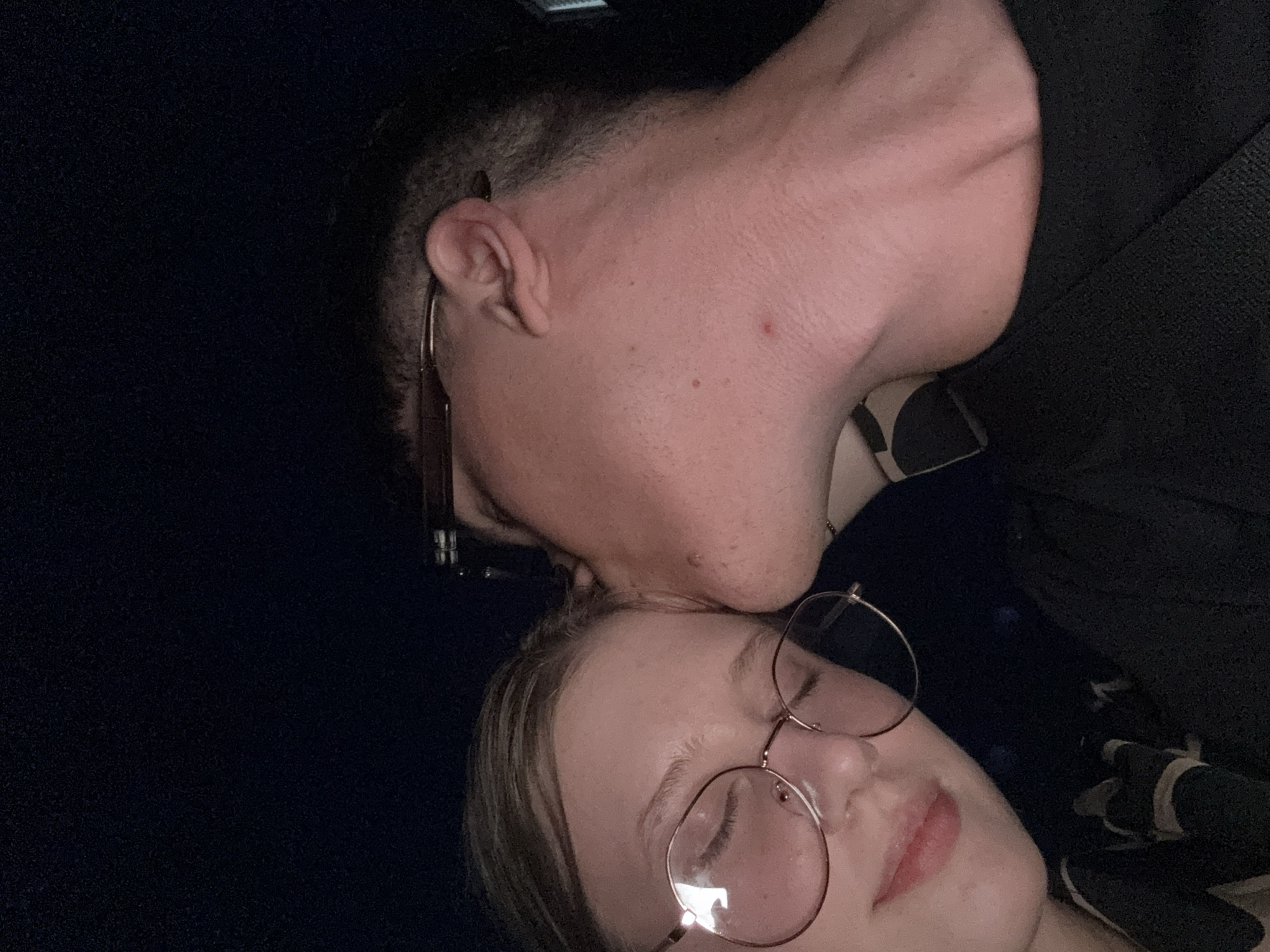 This is a close-up, indoor, eye-level, medium shot of a young Caucasian couple at night. The female subject is on the left, with her eyes closed and wearing round, wire-rimmed glasses. She has light brown hair and fair skin. Her lips are slightly parted, and she appears to be smiling faintly. The male subject is on the right, leaning in to kiss the female subject on her forehead. He has short, dark hair and is wearing glasses. The background is dark, suggesting an indoor setting at night. The image quality is moderate, with some graininess and low light. There is no indication of the city of Tiraspol, Moldova, within the image itself.