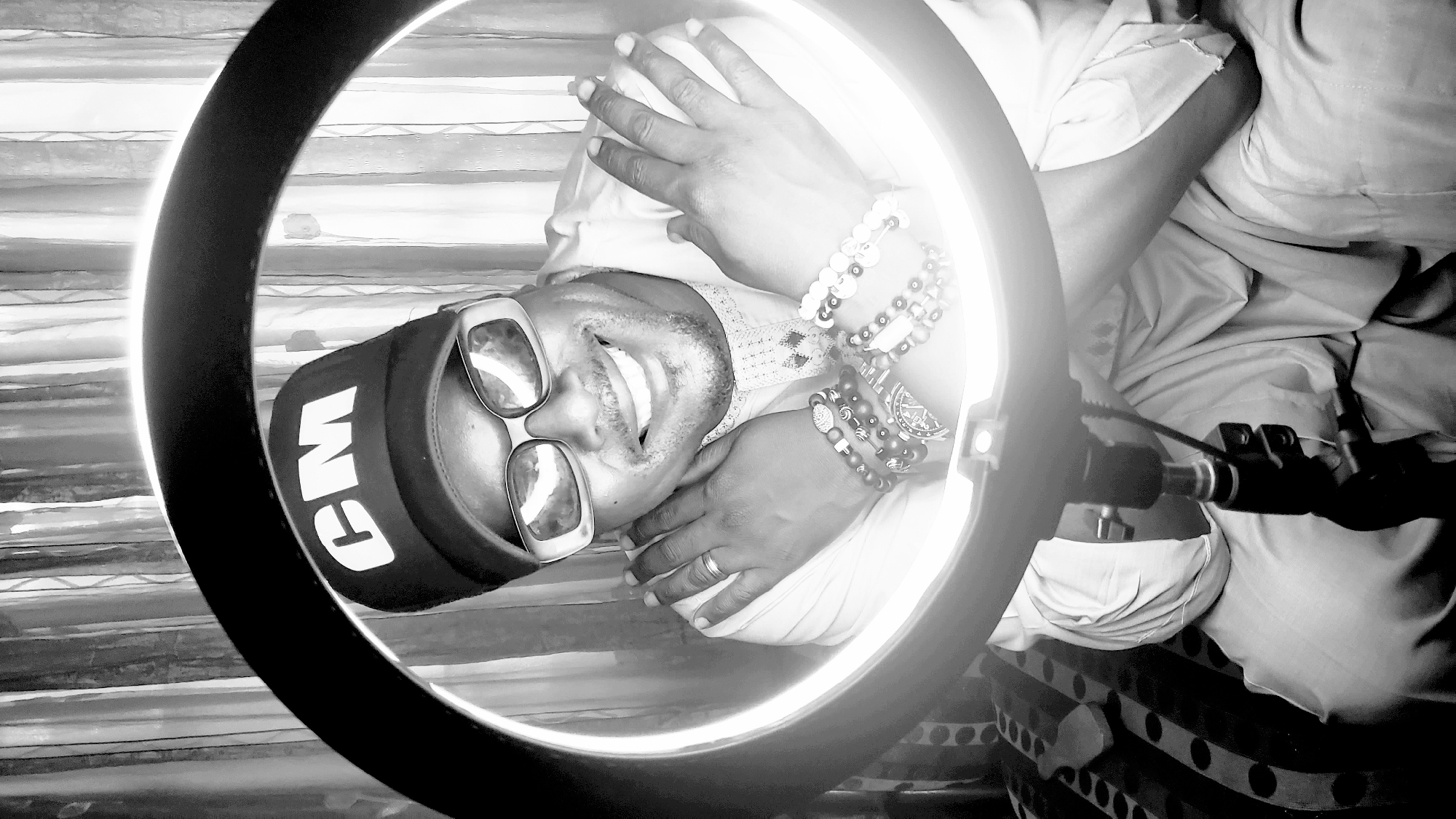 A black and white, low-angle selfie shows a man smiling with open eyes and mouth. He is wearing sunglasses with wide square frames and a black baseball cap with the letters "CM" in white block letters. He also wears several beaded bracelets on his left wrist. A ring light surrounds his head and shoulders, casting a bright light. The man appears to be lying on his back in bed. The bed sheets are white and partially wrinkled, and a patterned blanket or cover is visible beneath the sheets. The ring light is mounted on a stand visible to the lower right, partially obscured by the bed linens. The location in the city of Narayi, Nigeria, cannot be verified from the image.