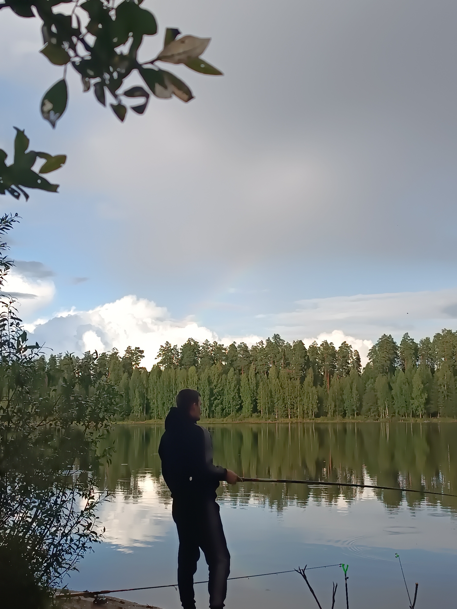 An adult male wearing a black hoodie and dark pants is fishing on the bank of a tranquil body of water, likely a lake, in Chaikovsky, Russia. He is holding a fishing rod, with another rod planted in the ground beside him. The water reflects the overcast sky and the dense green forest lining the opposite bank. The scene is partially obscured by the leaves of a tree in the foreground.