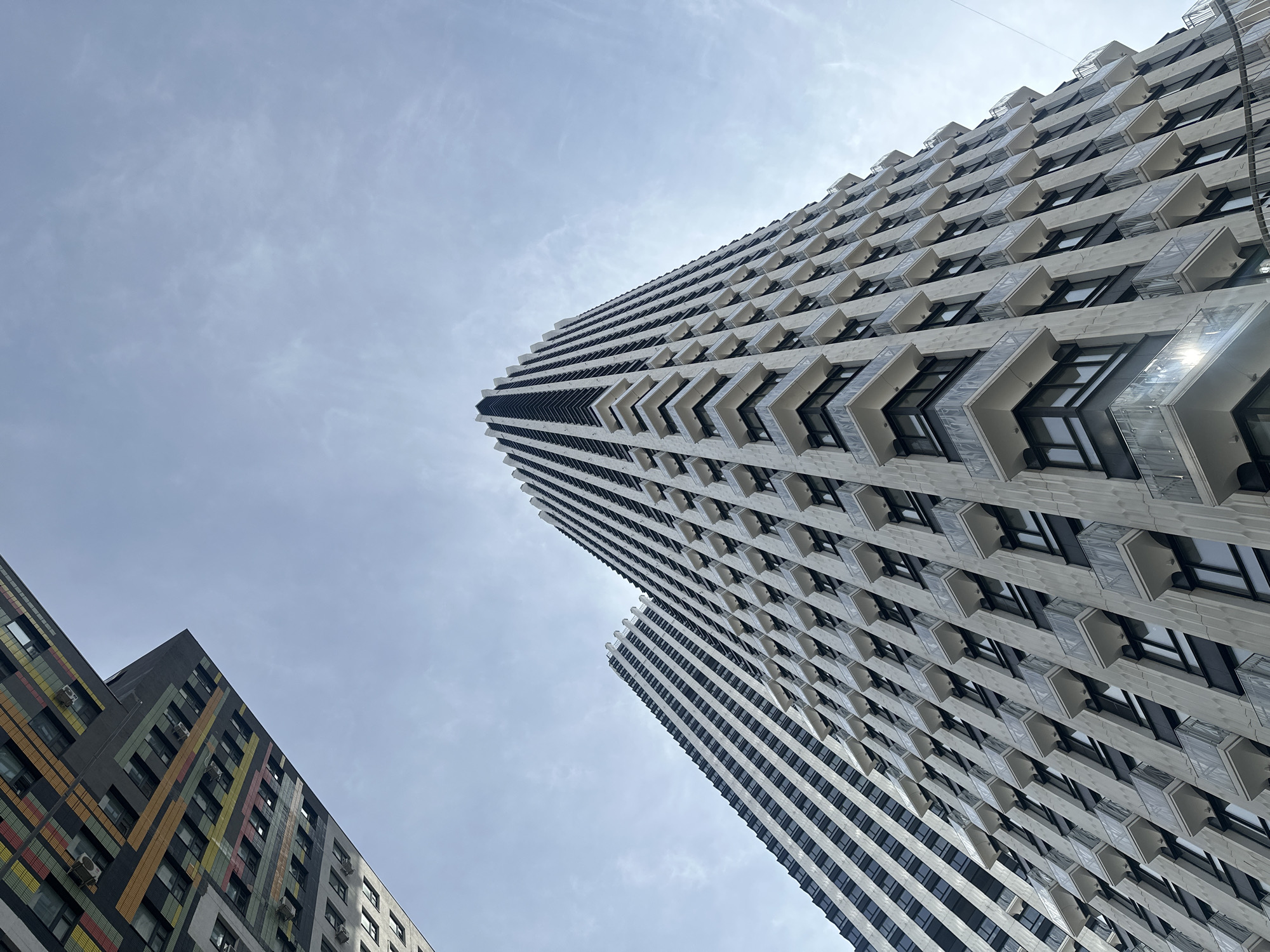 The image shows a low-angle view of modern architecture in Moscow, Russia. Two tall buildings dominate the frame, with the taller one on the right. This building has a distinctive facade with white and black horizontal stripes and protruding, chevron-shaped balconies. The second building, on the left, is shorter and features a facade with a gray base and colorful vertical stripes of yellow, red, and green. The sky is overcast, with thin clouds providing diffused light. The buildings appear to be residential apartment buildings based on the pattern of windows and balconies. There are no people visible in the image.